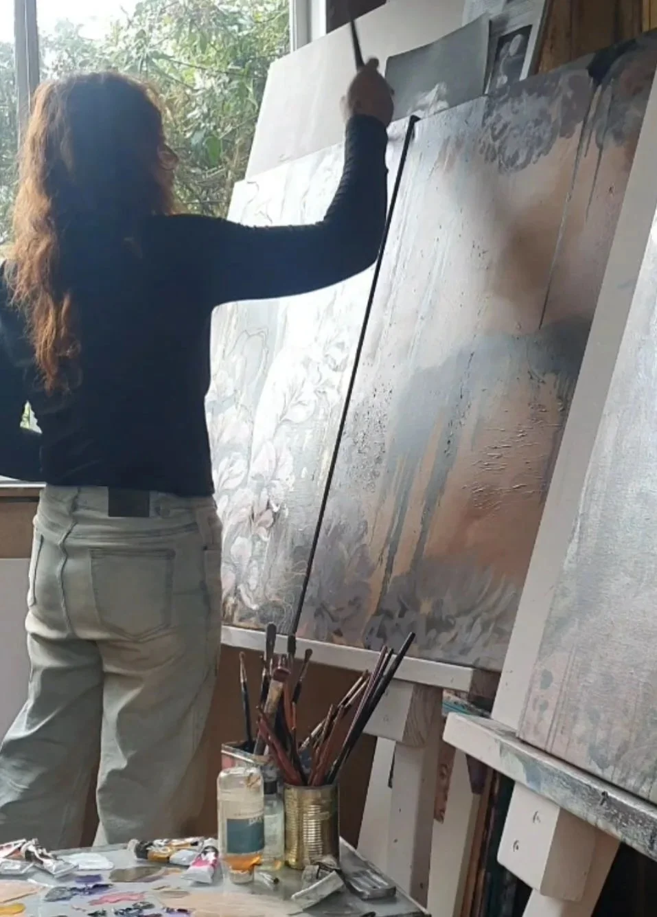 An artist with long curly hair painting on a large canvas in a studio, sunlight coming through a window, art supplies on a table in the foreground.