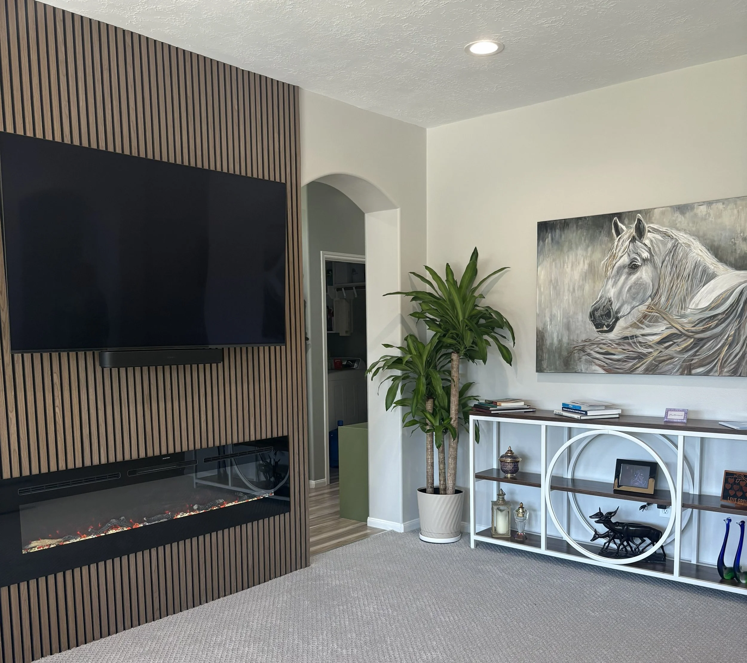 A living room corner featuring a black fireplace with a wooden slat wall on the left, a large flat-screen TV, and decorative items including a plant, books, and artwork on a white shelved unit. A painting of a white horse hangs on the white wall abov