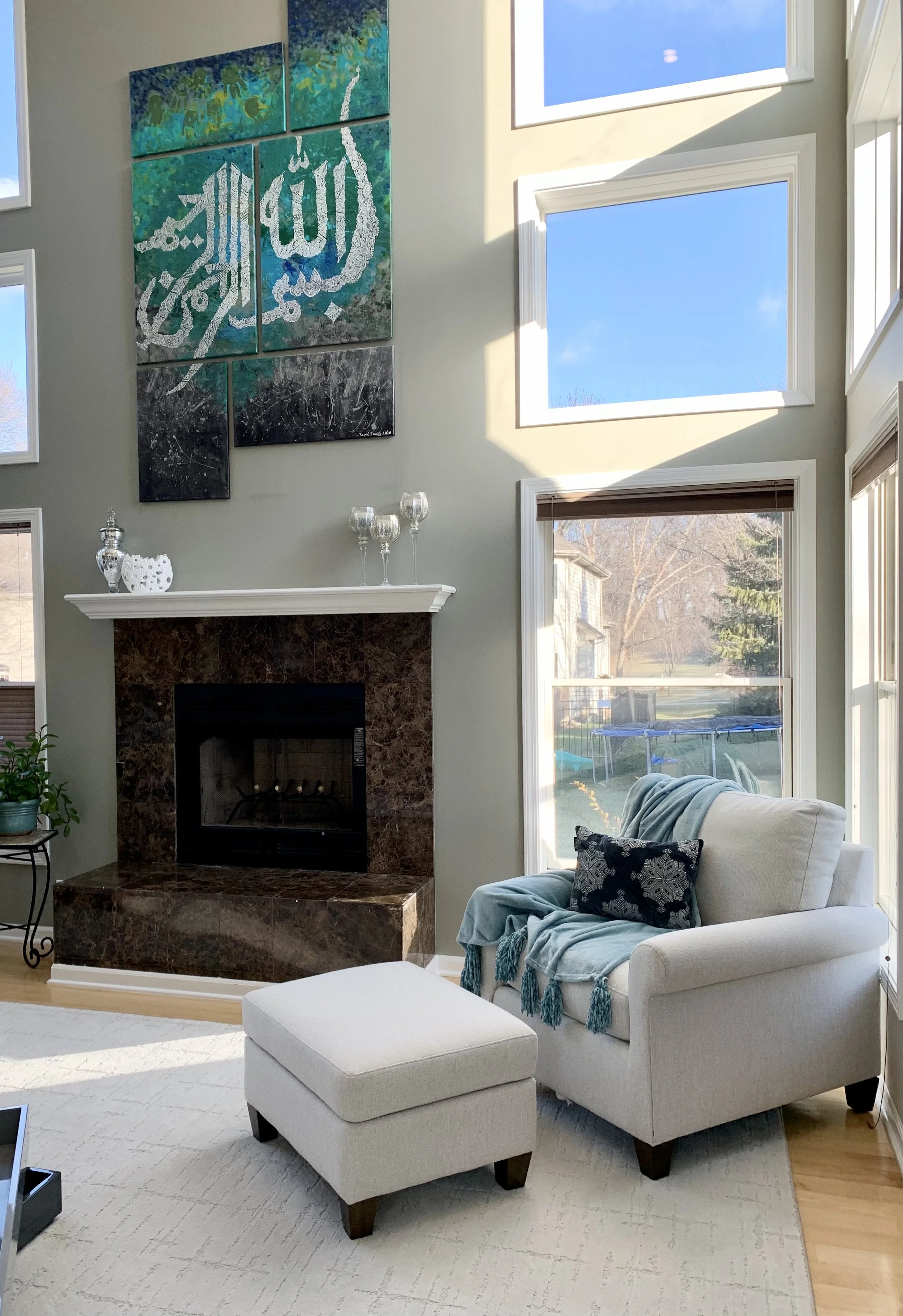 A living room with a white armchair and matching ottoman, a marble fireplace, and a wall hanging featuring Arabic calligraphy. Large windows show a backyard with a trampoline and trees, and sunlight illuminates the space.