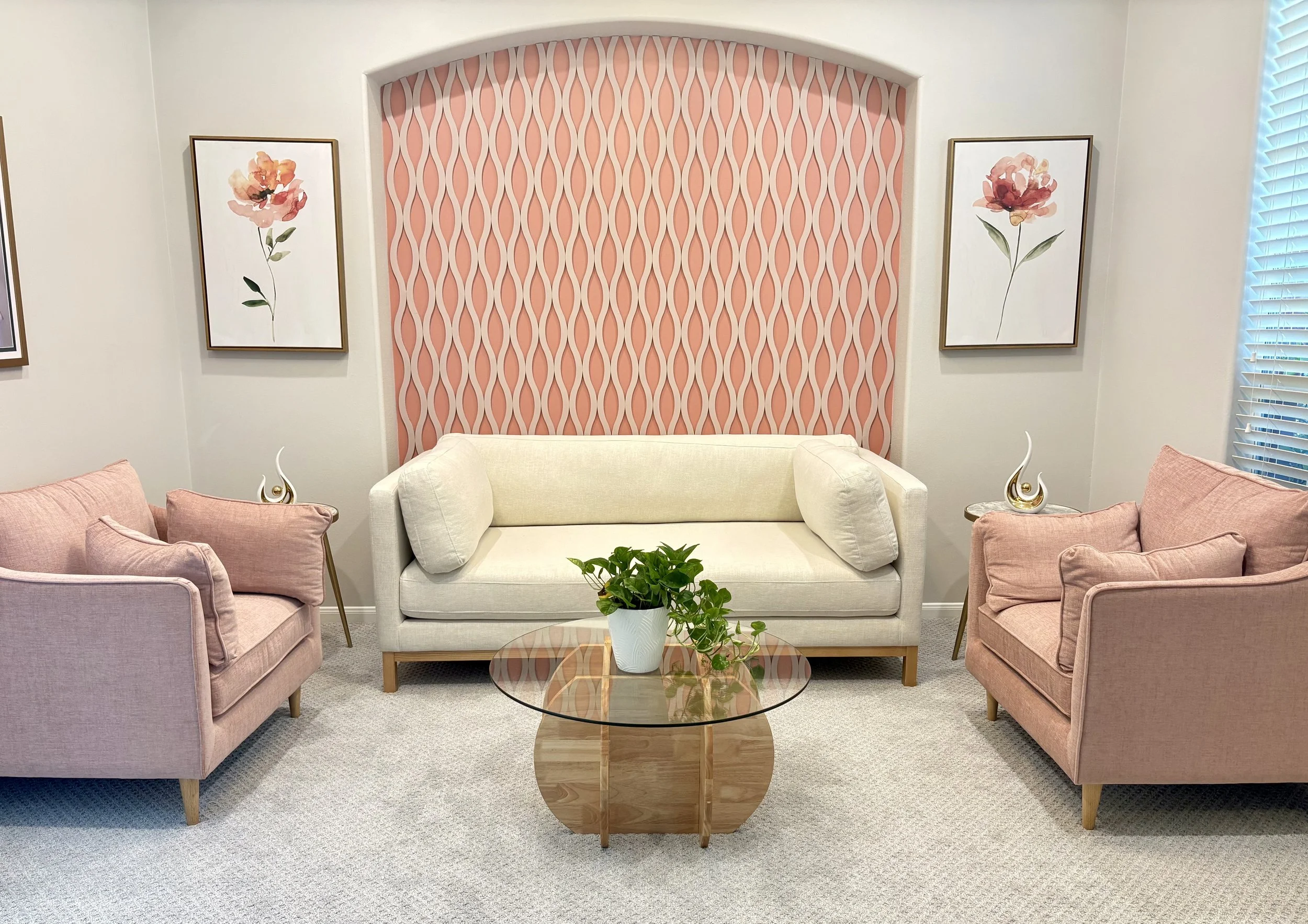 Living room with two pink armchairs, a white loveseat, a round glass coffee table with a potted plant, pink geometric patterned accent wall, and framed floral artwork.