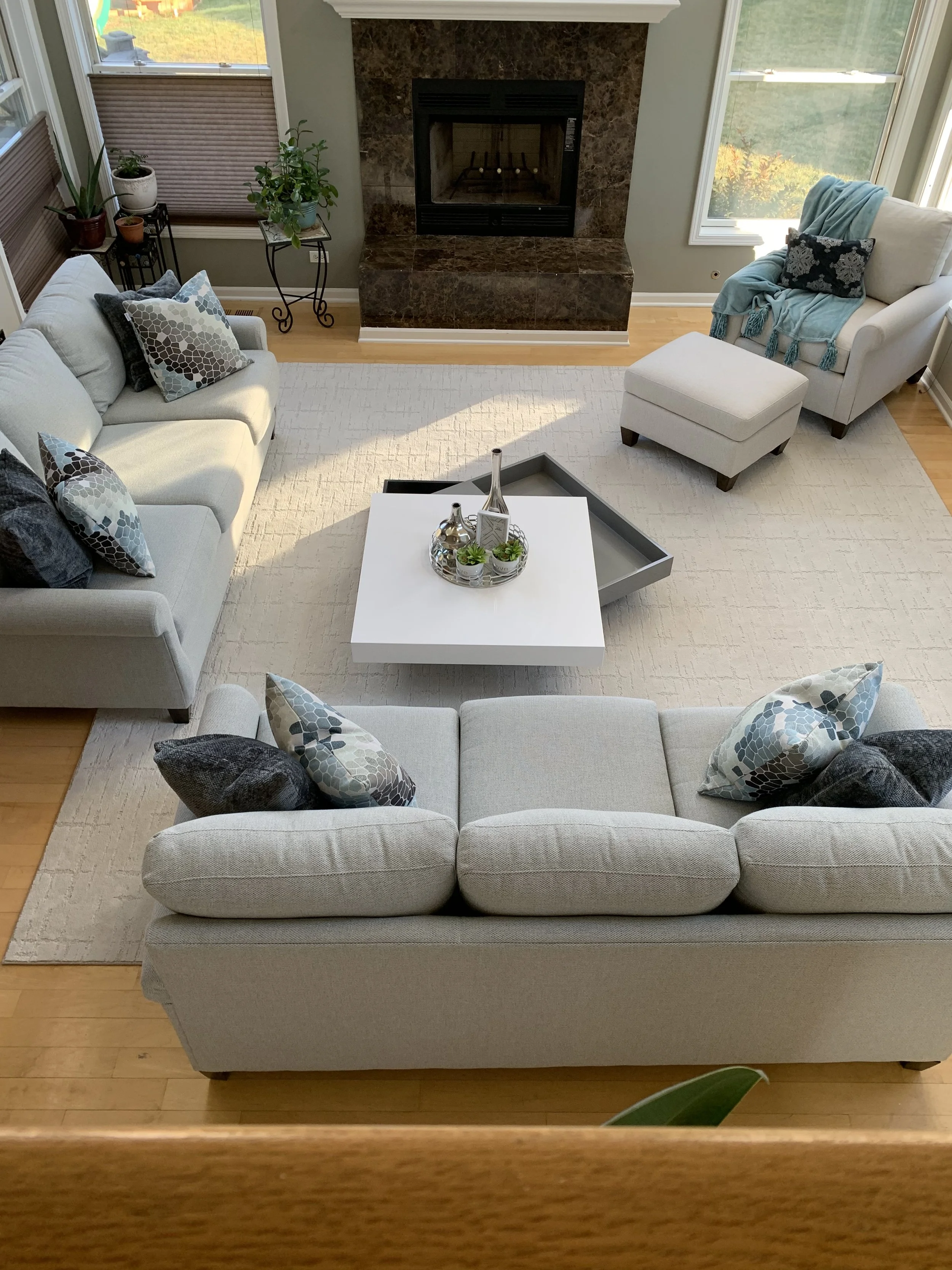 Living room with a white sofa, a white armchair with a blue blanket, plants near the window, a fireplace with a brown marble surround, a white coffee table with decorative vases and succulents, and large windows showing an outdoor garden.
