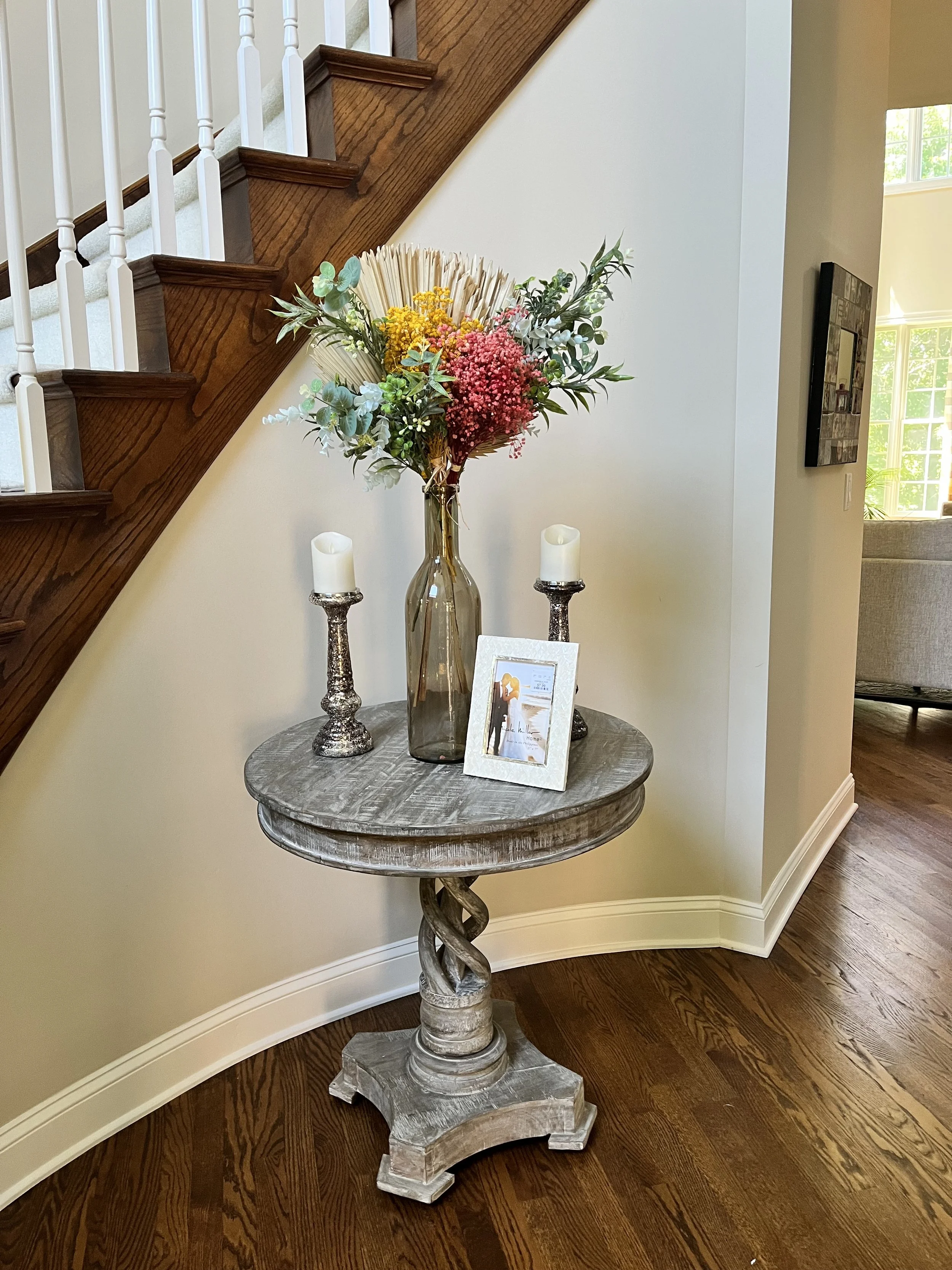 Elegant round wooden table featuring a large glass vase with a colorful floral arrangement, flanked by two silver candlesticks with white candles, in a well-lit room with hardwood floors and staircase.
