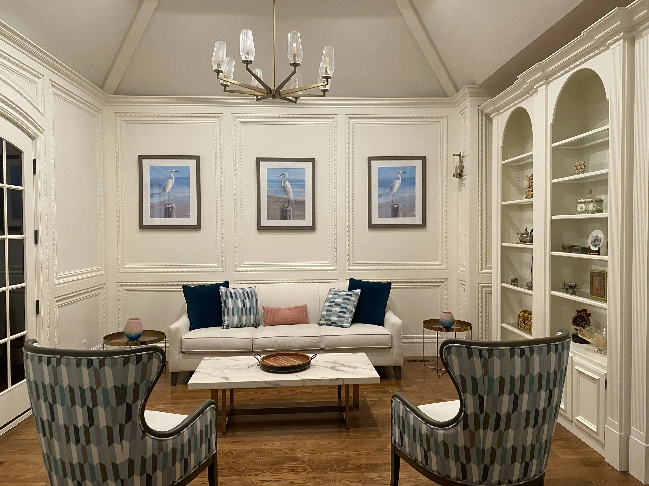 Living room with white walls, three framed bird paintings, a white sofa with patterned pillows, two small round side tables, and six dining chairs with blue, gray, and white geometric fabric facing a marble-topped coffee table, illuminated by a moder