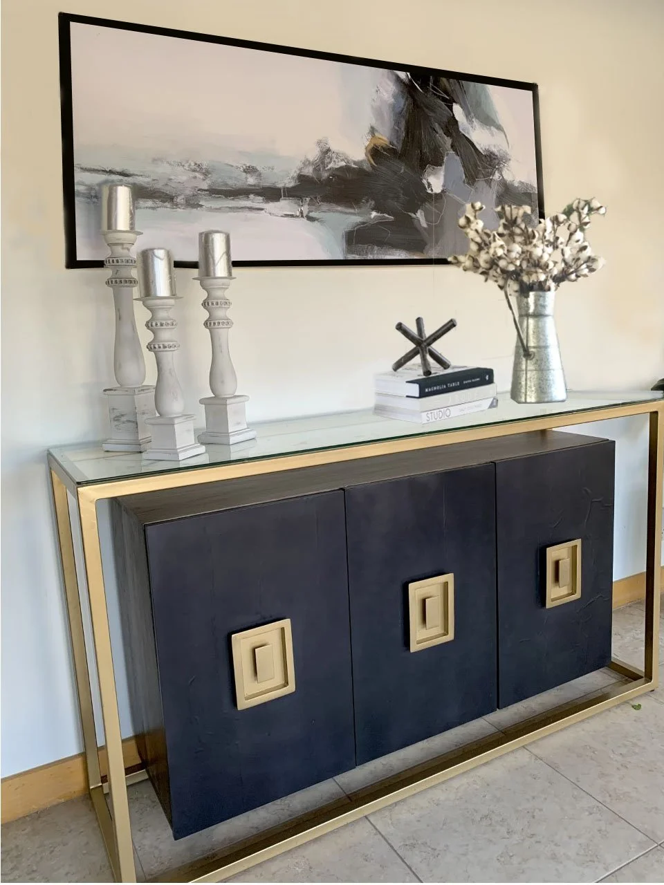A black and gold sideboard with a glass top, decorated with white candlesticks, a stack of books, a metallic vase with cotton branches, and a black abstract artwork hanging above.