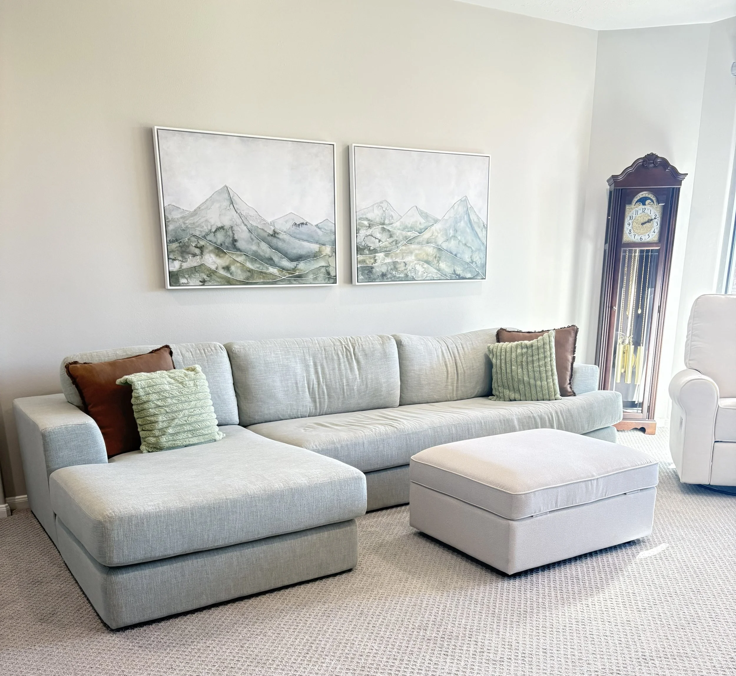 Living room with white upholstered sofa, brown and green throw pillows, two separate wall art pieces depicting mountain landscapes, and a wooden grandfather clock in the corner.