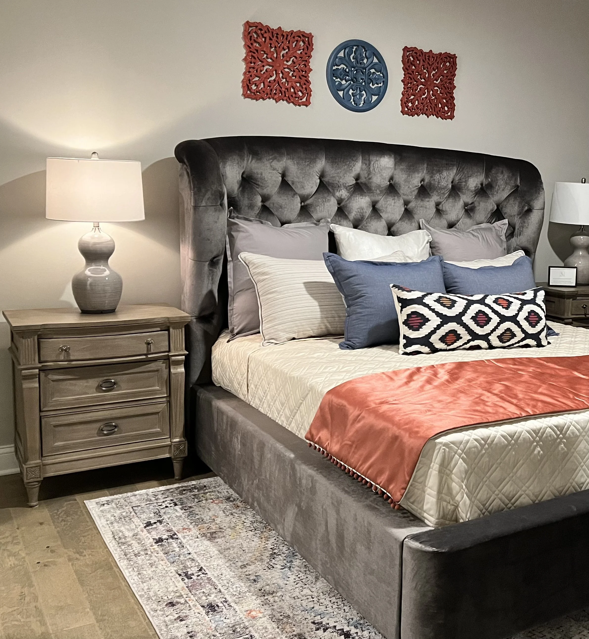 Bedroom with a taupe tufted velvet headboard, a wooden nightstand with three drawers and metal handles, two table lamps with white shades, various pillows on the bed, and wall art including a red lattice design, a blue circular decorative piece, and 