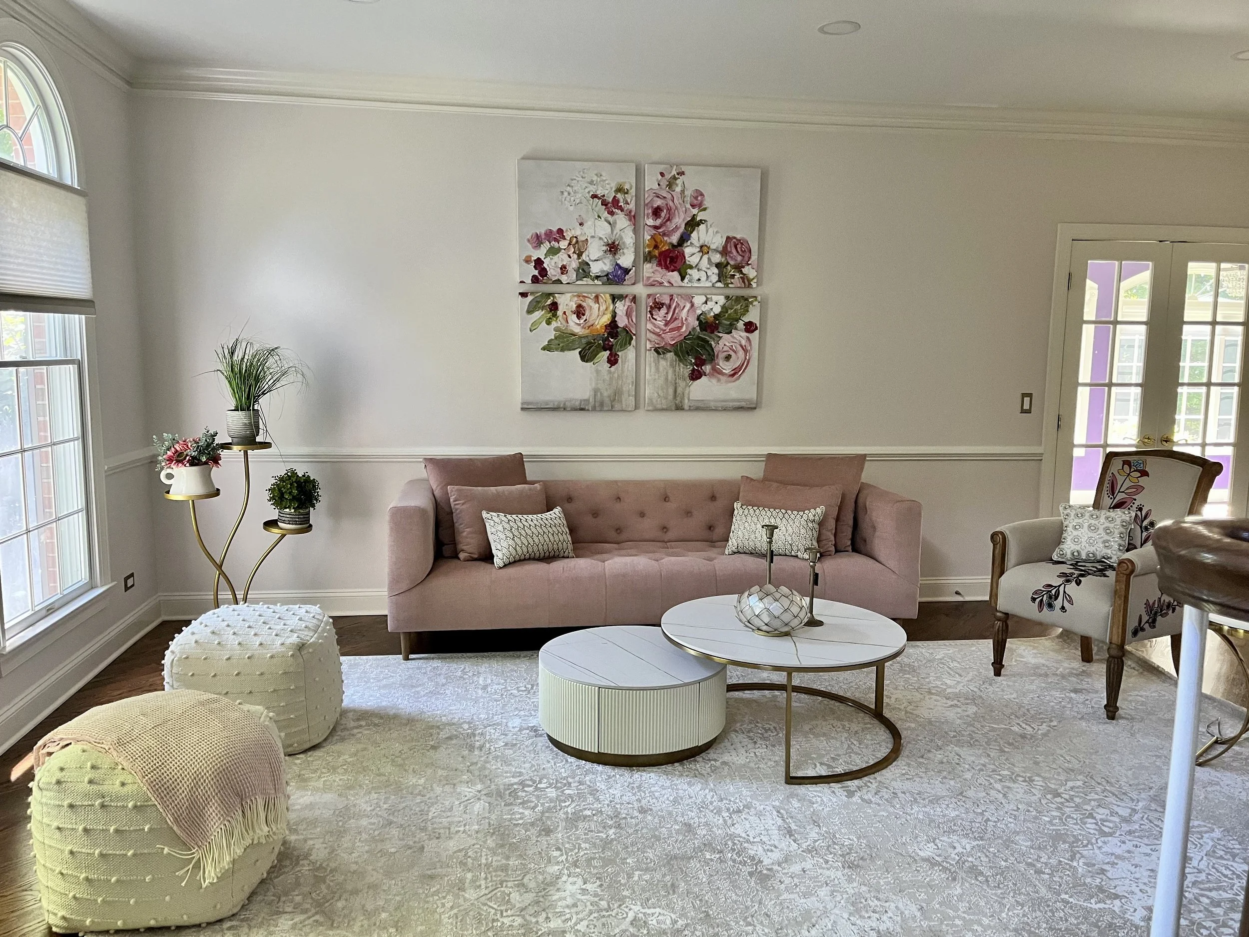 Living room with pink sofa, floral wall art, white coffee tables, wicker ottomans, and potted plants near large windows.