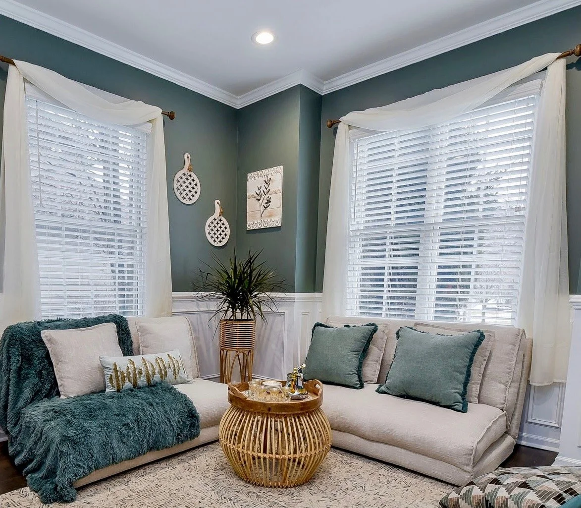 Living room with teal walls, white trim, large windows with white blinds and sheer curtains, beige sofa with teal and beige pillows, and a round wooden coffee table with a plant and tray. Wall decor includes two white decorative pieces and a framed a