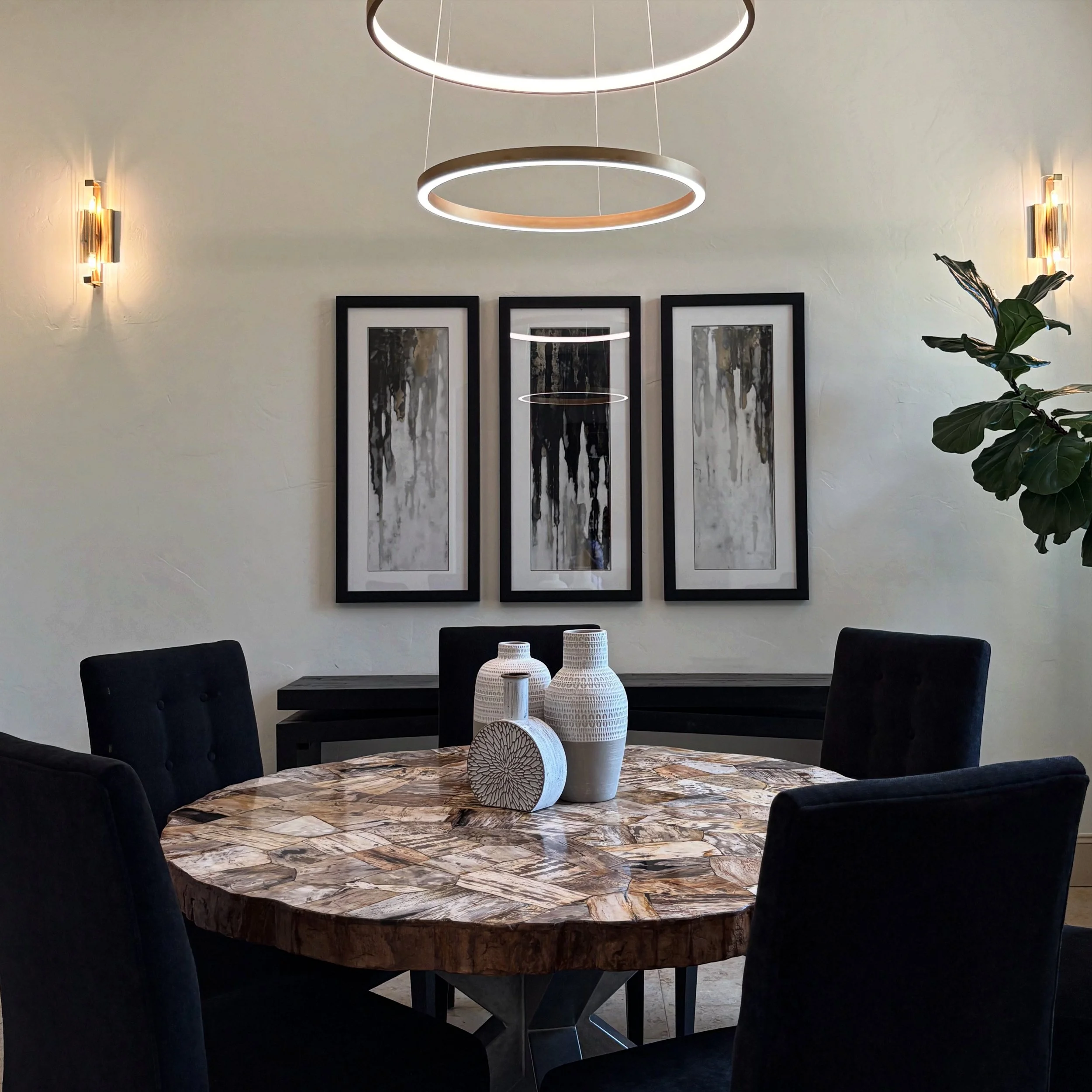 Modern dining room with a marbled wooden table, four black chairs, three white decorative vases, abstract framed artwork on the wall, and contemporary lighting fixtures including a chandelier and wall sconces.