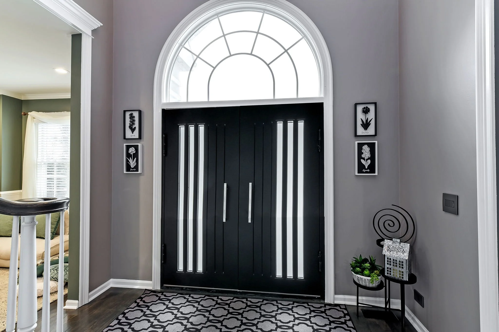Entryway with black front door, decorative side panels, semi-circular window, gray walls, black and white patterned rug, framed floral artwork, and small plants on a black side table.