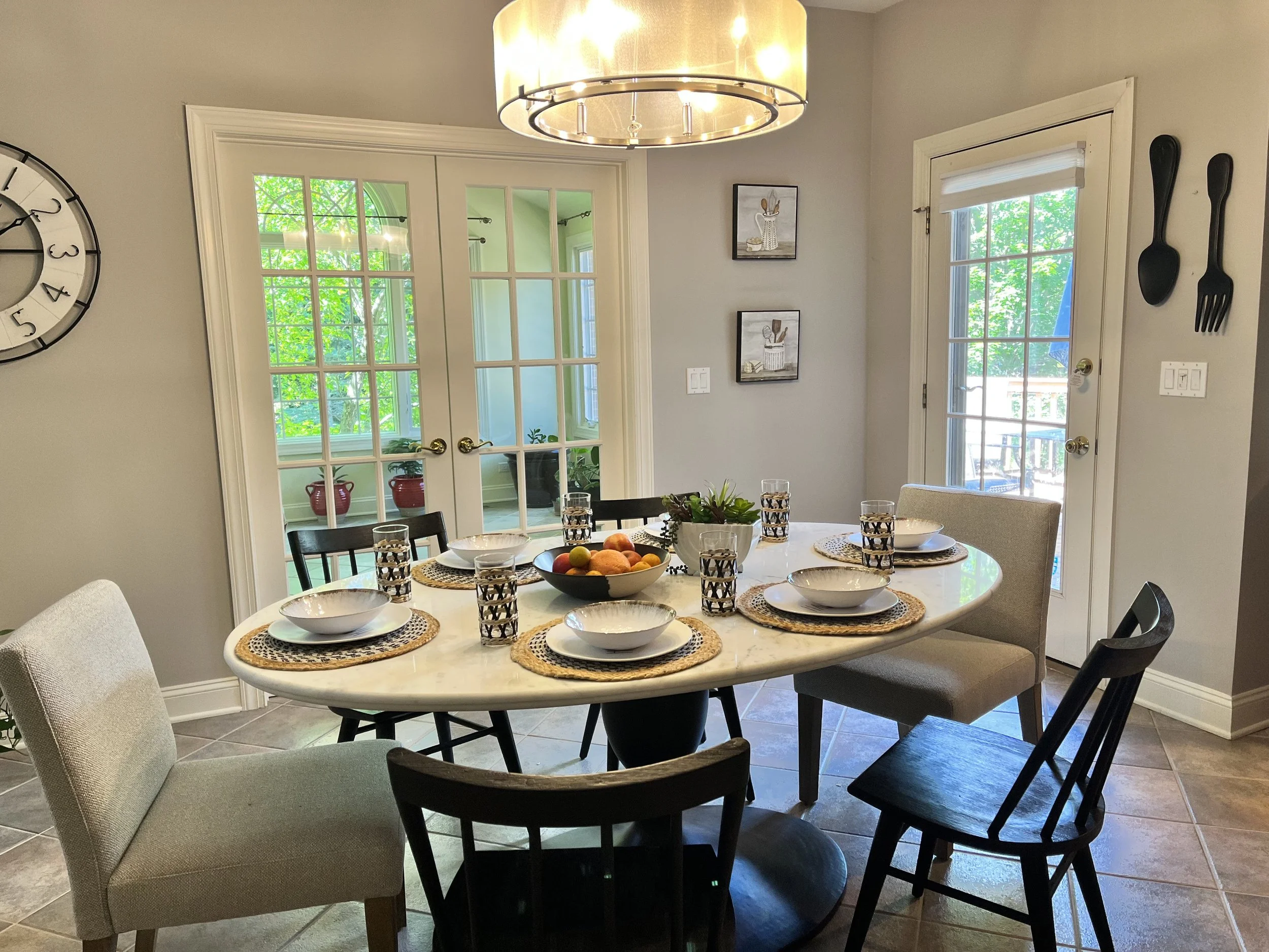A dining room with a round table set with plates, glasses, napkins, and a centerpiece of fruit. The table is surrounded by eight chairs of different styles. The room has two sets of glass-paneled double doors leading to an outside porch, with greener
