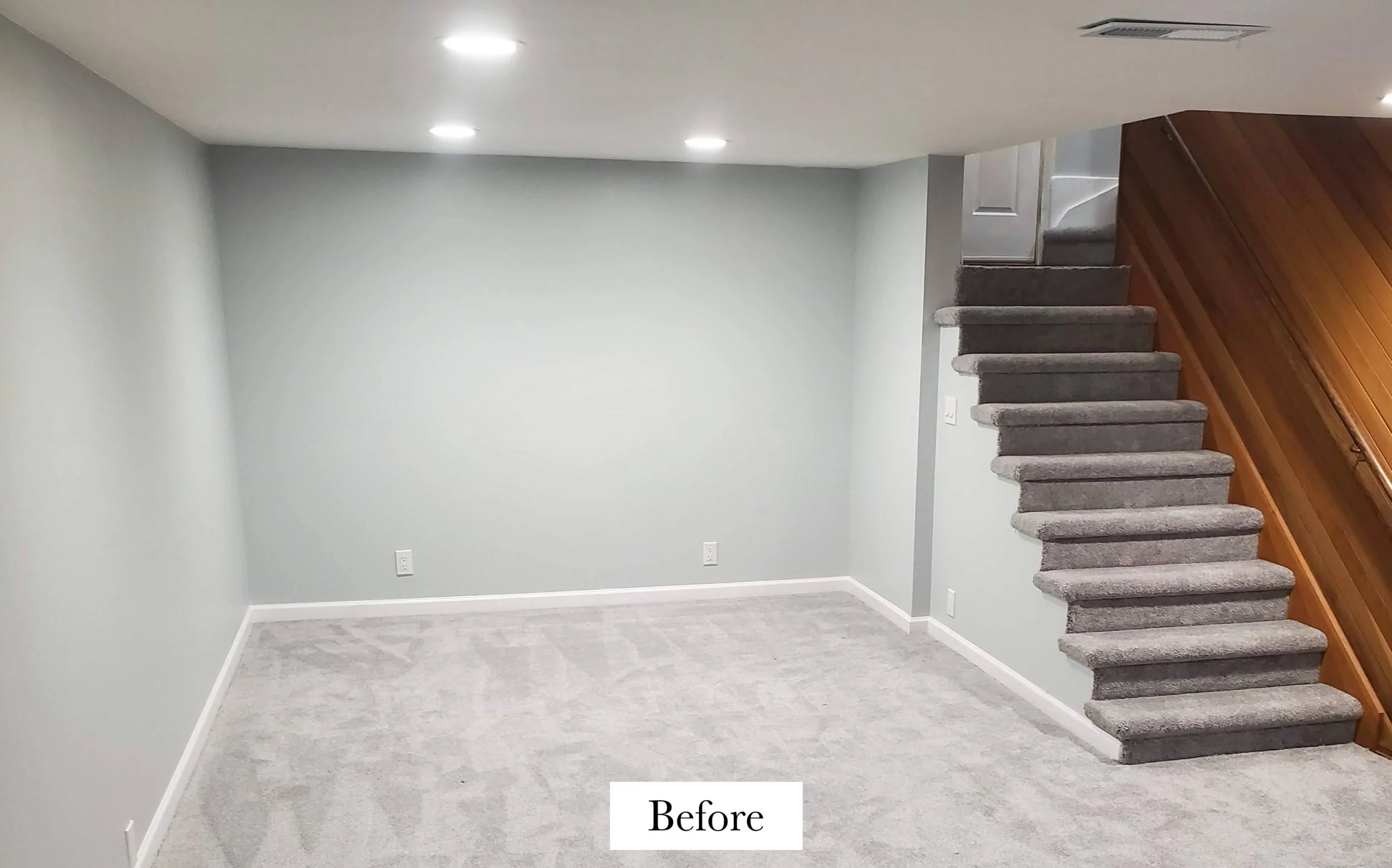 An empty room with light gray walls, light gray carpet, and a staircase with gray carpeted steps leading to an upper level. The ceiling has recessed lighting.