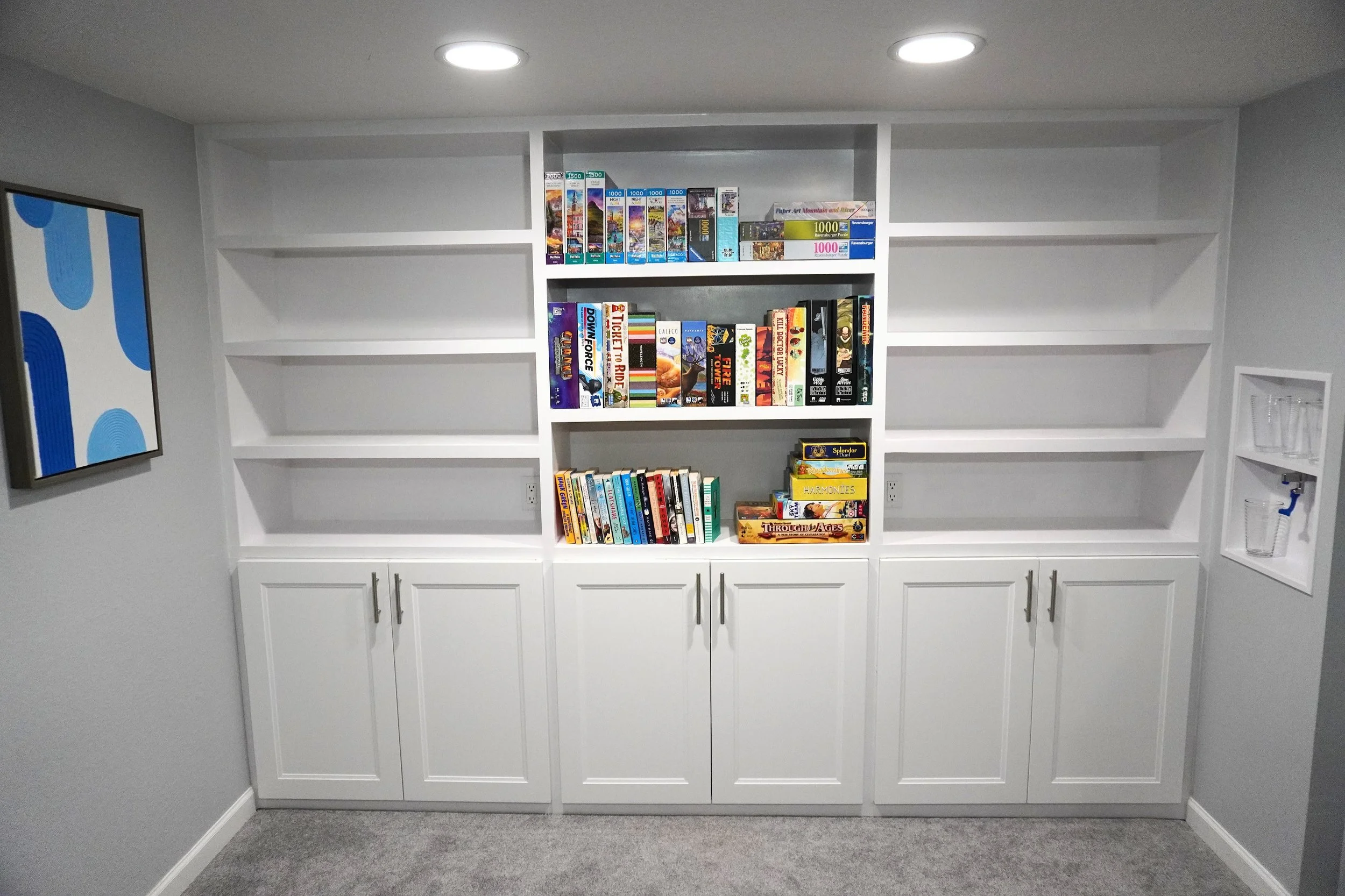White built-in bookshelf filled with board games and books in a room with gray walls and carpet, featuring a colorful abstract painting on the left wall and a small white shelf with glasses and a water faucet on the right wall.