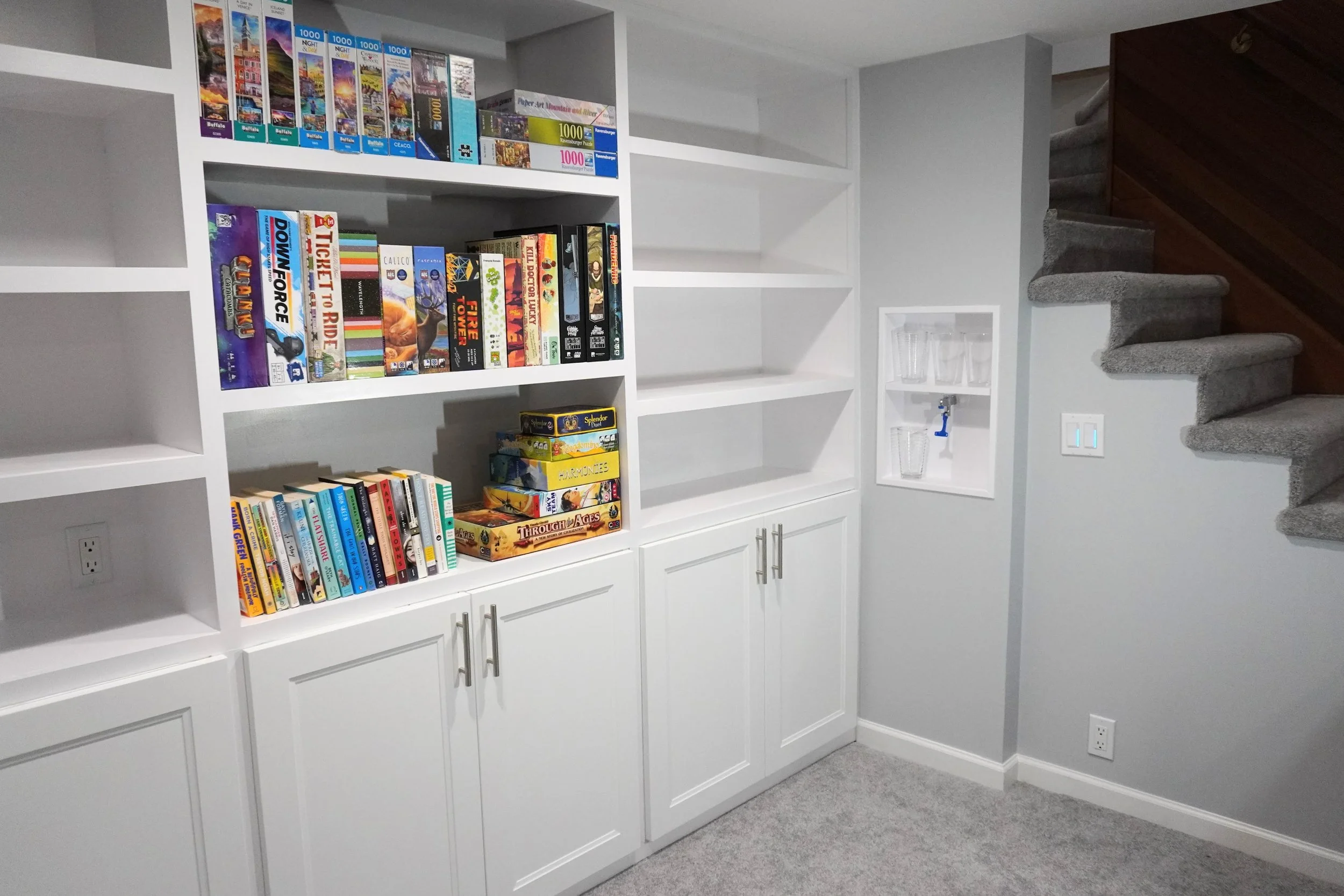 White built-in shelves filled with books and board games in a room with gray carpet and wall, stairway with gray carpet and wooden railing in background.