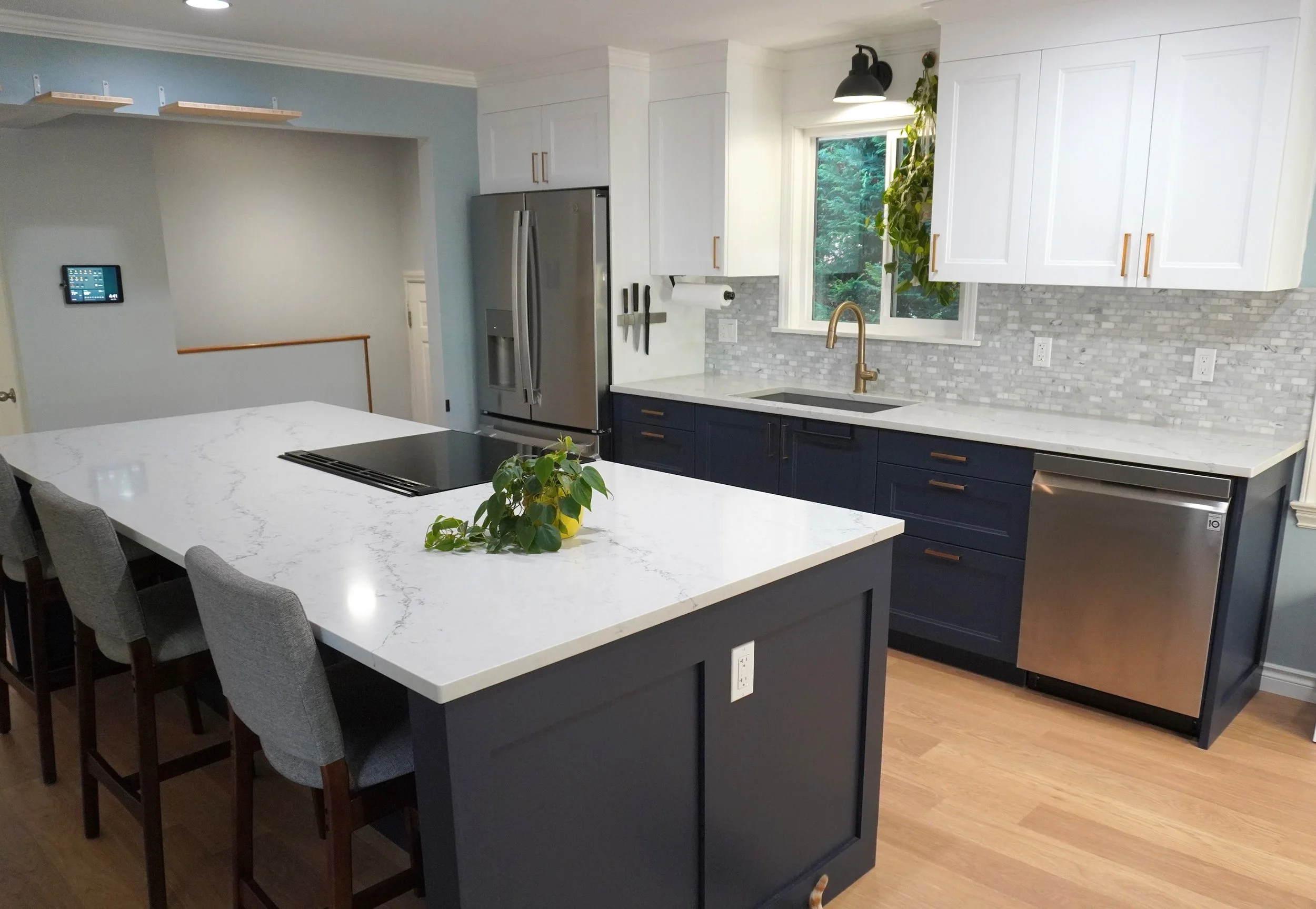 Modern kitchen with white and navy blue cabinets, stainless steel appliances, and a white marble countertop island with seating. There are hanging knives, a window with greenery outside, and a small plant on the island.
