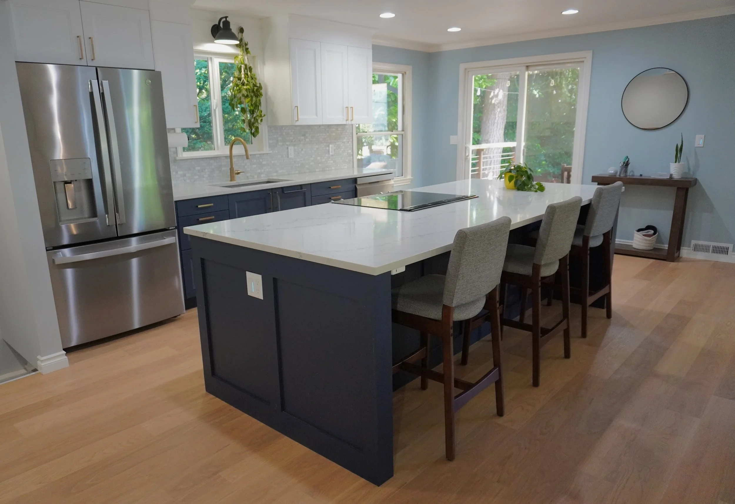 Modern kitchen with blue and white cabinetry, stainless steel refrigerator, marble island with seating, hardwood flooring, and a sliding glass door leading to an outdoor space. Features include a plant on the island, a mirror on the wall, and a small