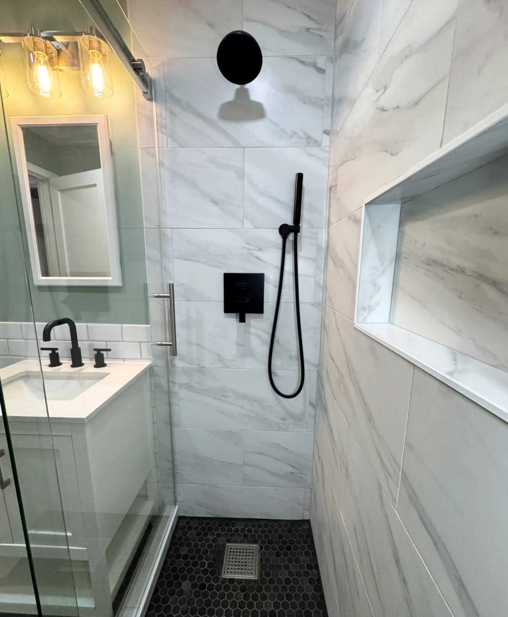 Modern shower area with black fixtures, a small built-in shelf, and a glass partition in a bathroom with white marble walls and a black hexagonal tile floor.