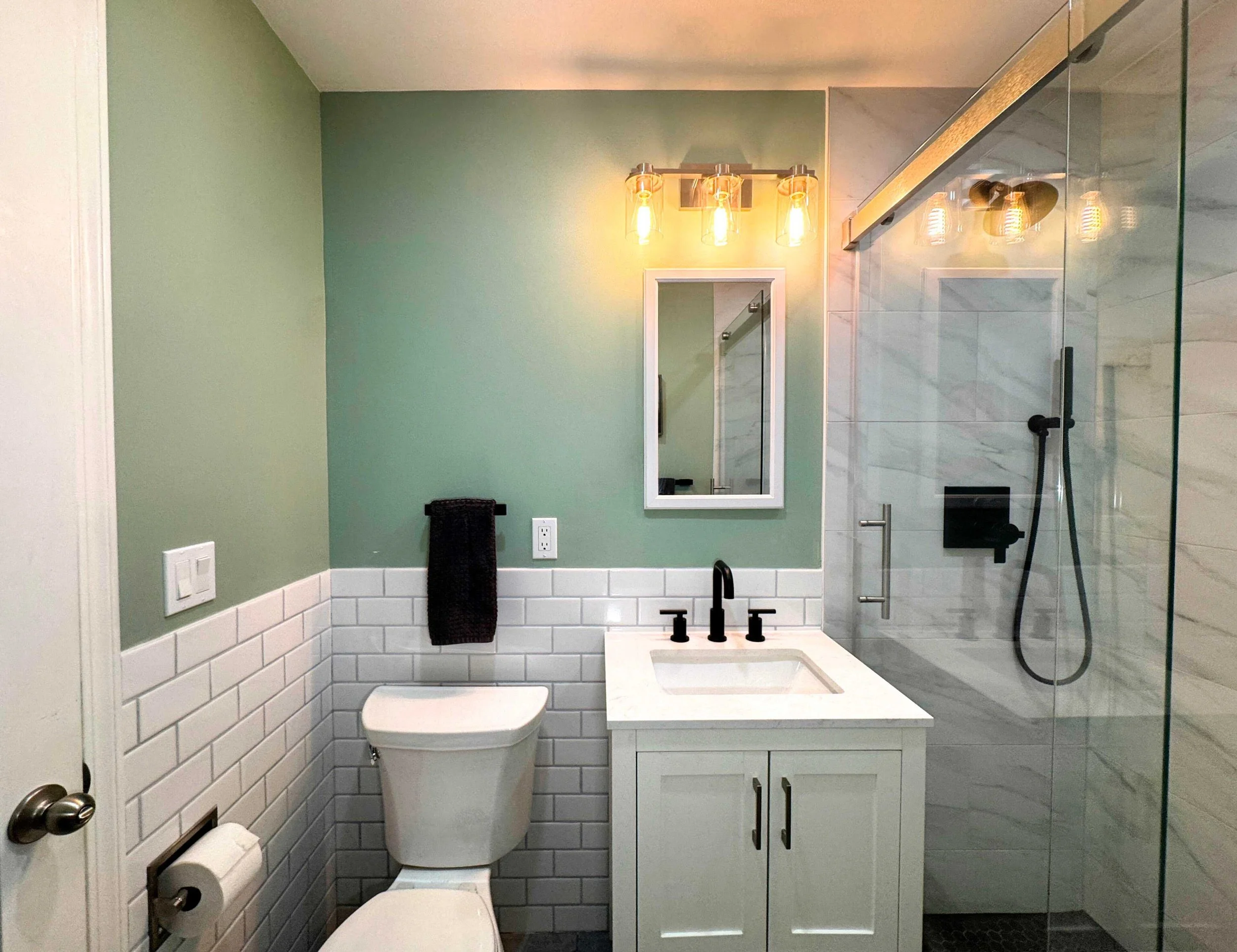 A modern bathroom with green walls, a white toilet, a small white vanity with a sink and black fixtures, a mirror, and a glass-enclosed shower with white marble tiles and a black handheld showerhead.