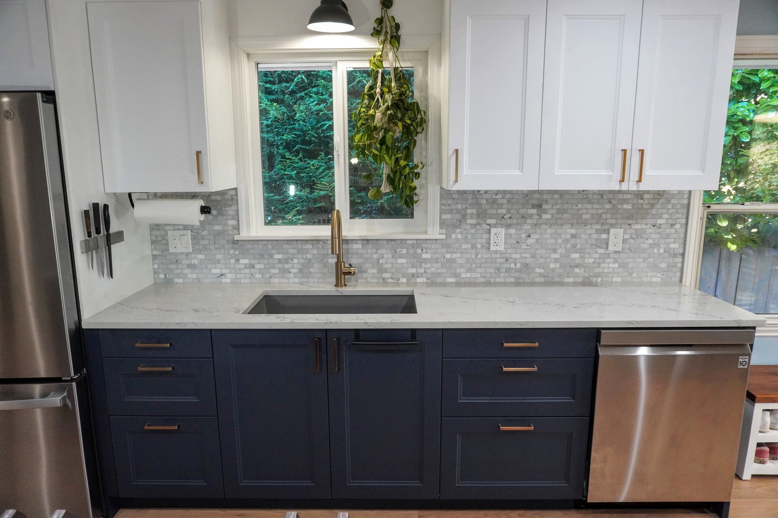 Modern kitchen with white upper cabinets, navy blue lower cabinets, a white marble countertop, and a gray tile backsplash. A sink with a gold faucet is positioned under a window showing green outdoor foliage. There are kitchen knives on a magnetic strip on the left wall and a hanging plant in the window.