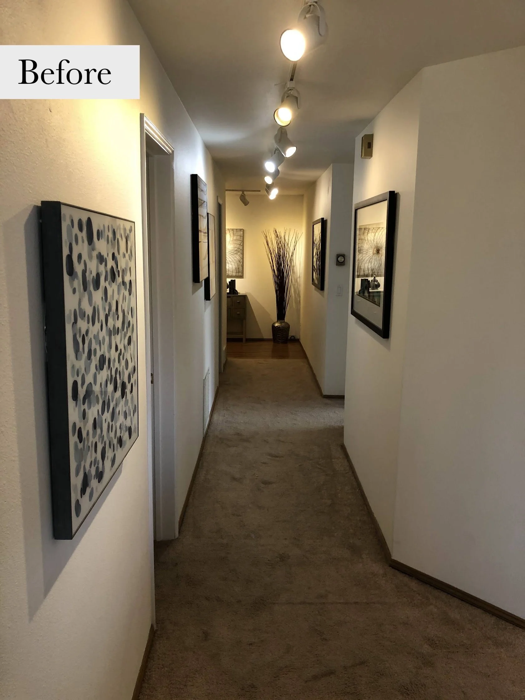 Long hallway with ceiling track lighting, beige carpet, and wall art, leading to a small table and decorative vase in the background.