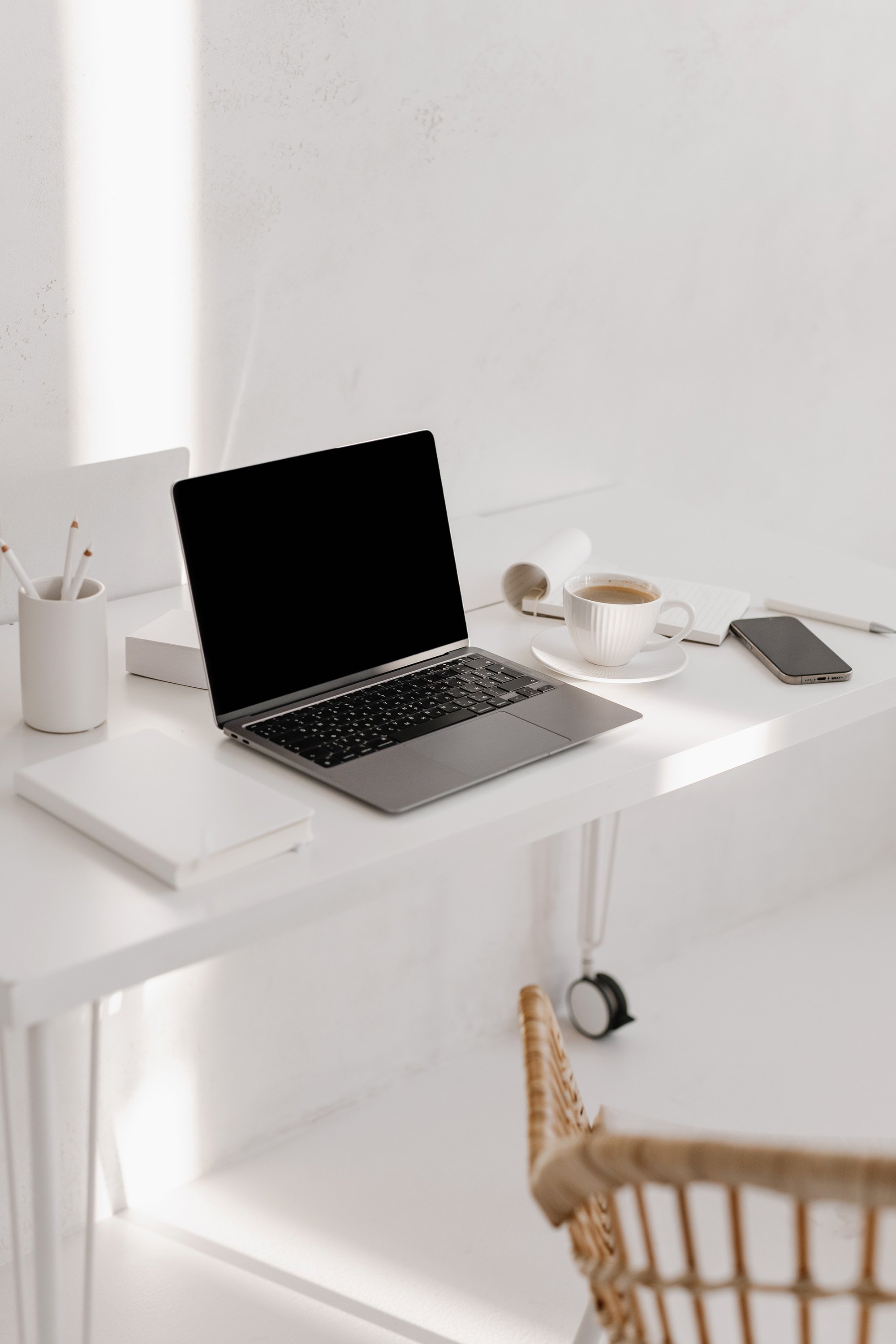 Minimalist white desk with a laptop, a cup of coffee, a smartphone, a notepad, and some stationery in a bright, modern workspace.
