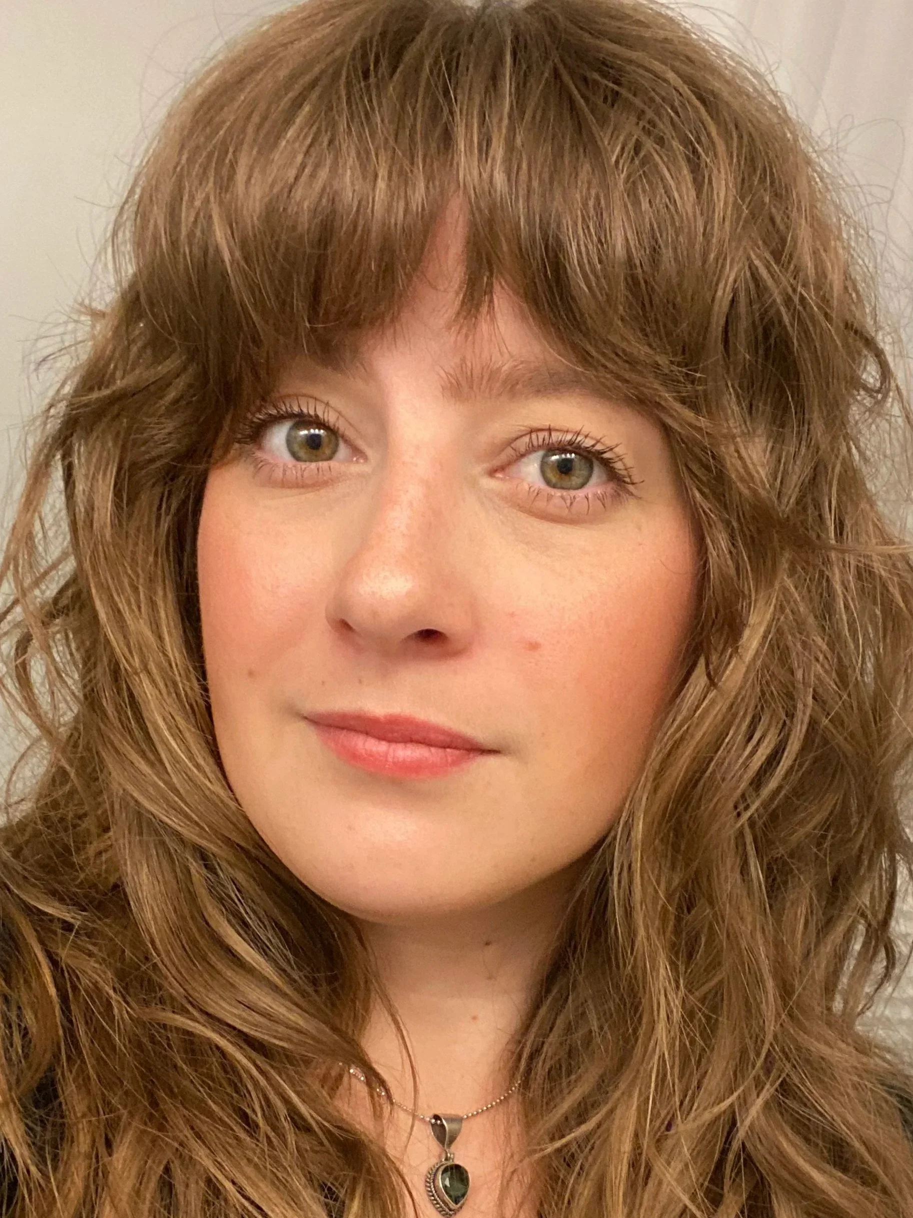 Close-up of a young woman with long, curly brown hair, fair skin, hazel eyes, and a gentle smile, wearing a heart-shaped pendant necklace.
