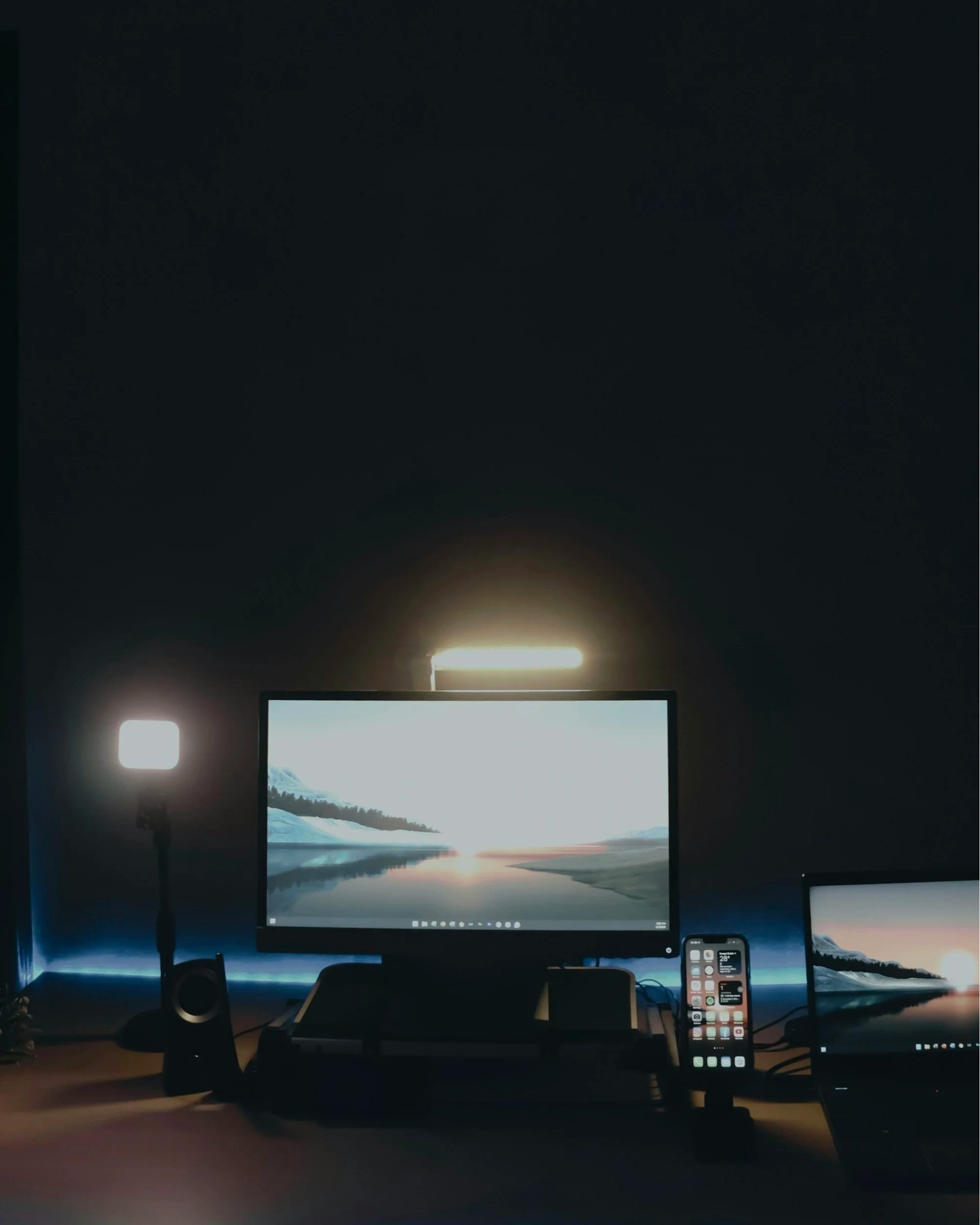 Computer desk setup with two monitors displaying a scenic landscape of mountains and water at sunset, with a smartphone showing app icons and a keyboard in front of the monitors, all in a dimly lit room.