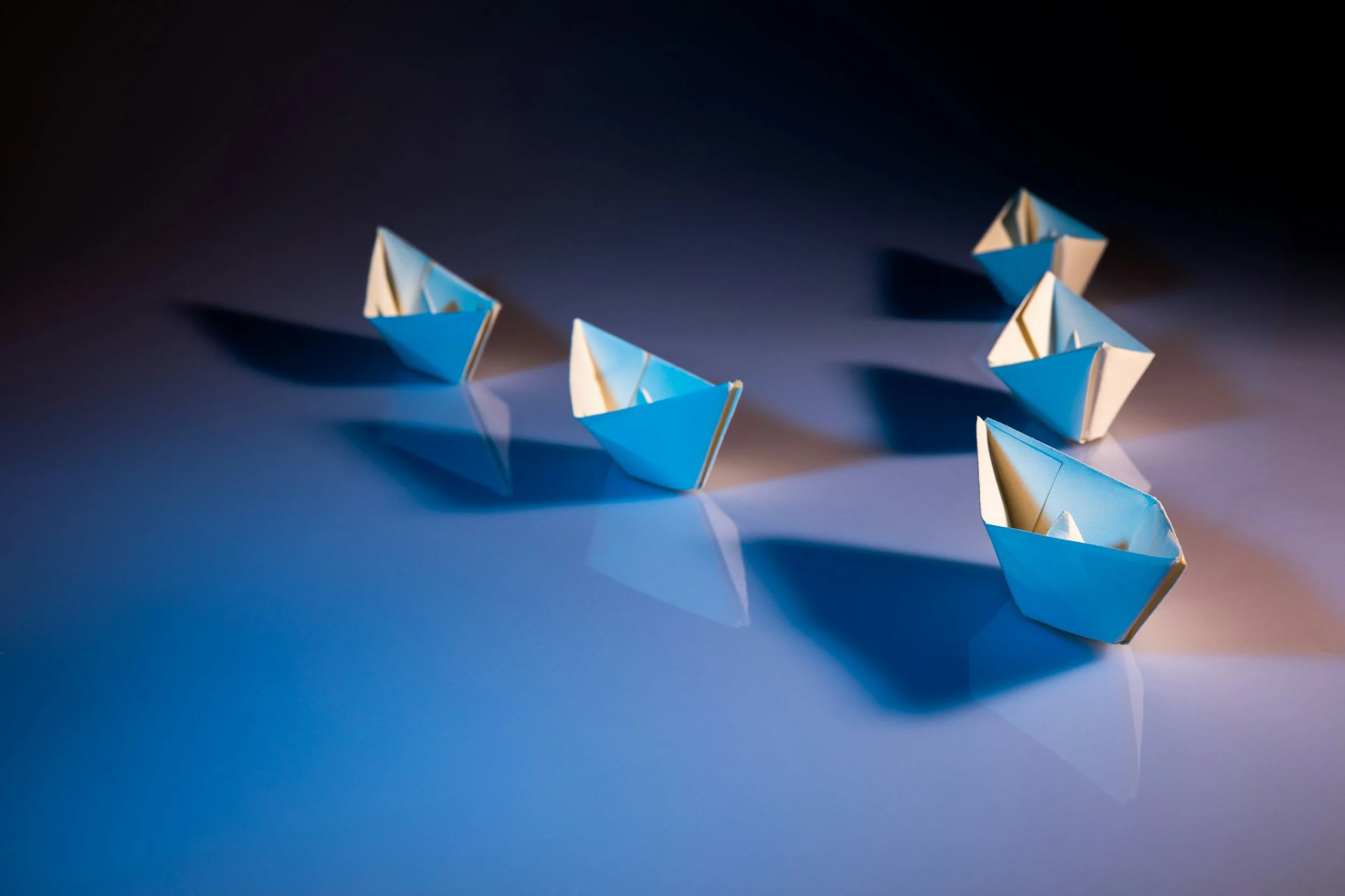 Multiple white and blue paper boats arranged on a dark and light patchy background, casting shadows.