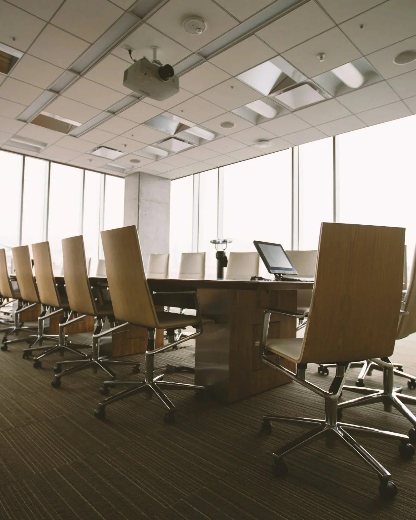 Empty modern conference room with a large wooden table, beige chairs, a digital conference phone, and a tablet. Floor-to-ceiling windows let in natural light, with ceiling-mounted projector visible.