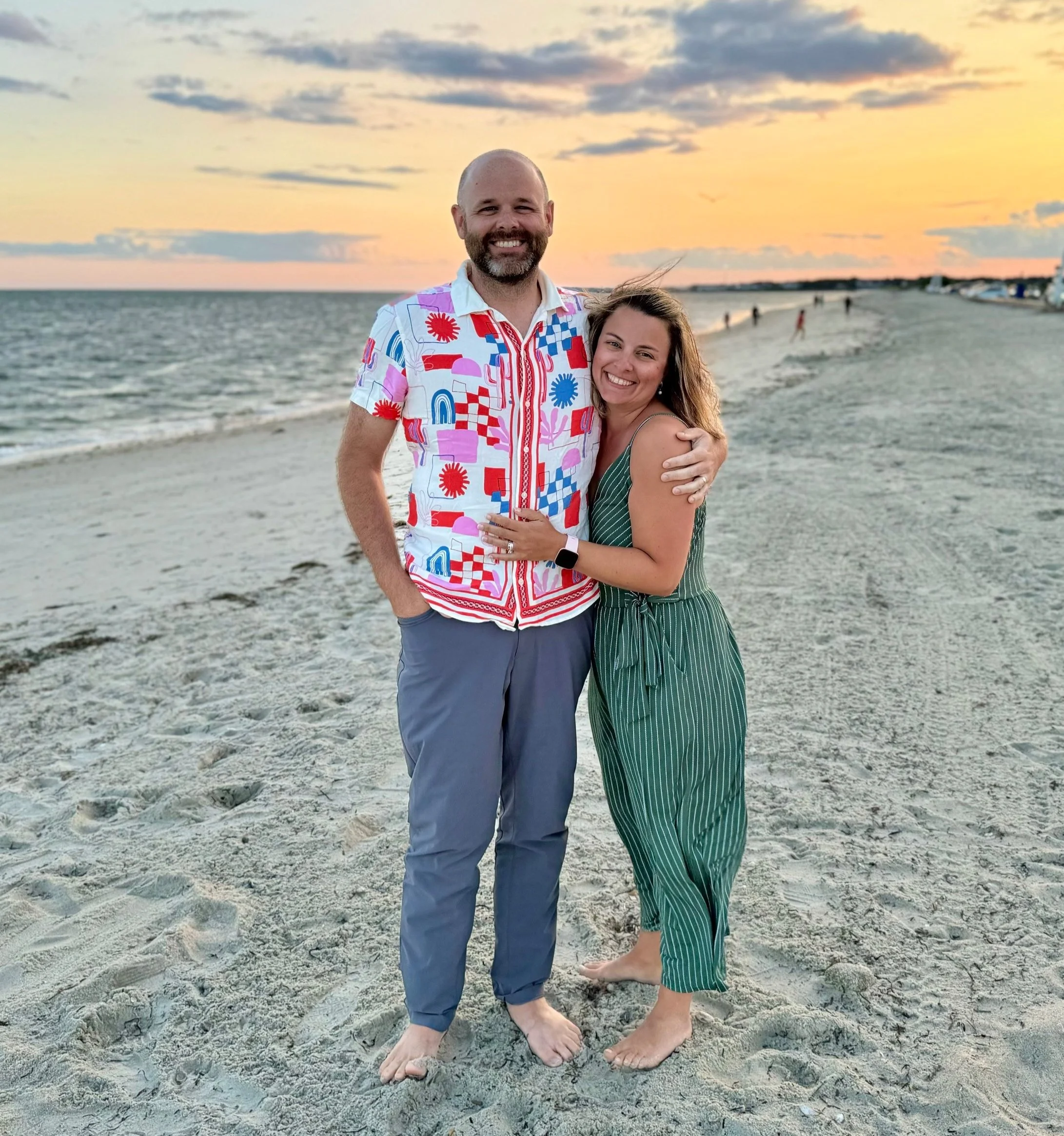 A smiling couple standing on a sandy beach at sunset, hugging each other. The man is wearing a colorful patterned shirt and gray pants, and the woman is in a green striped jumpsuit. The ocean and a sunset sky are in the background.