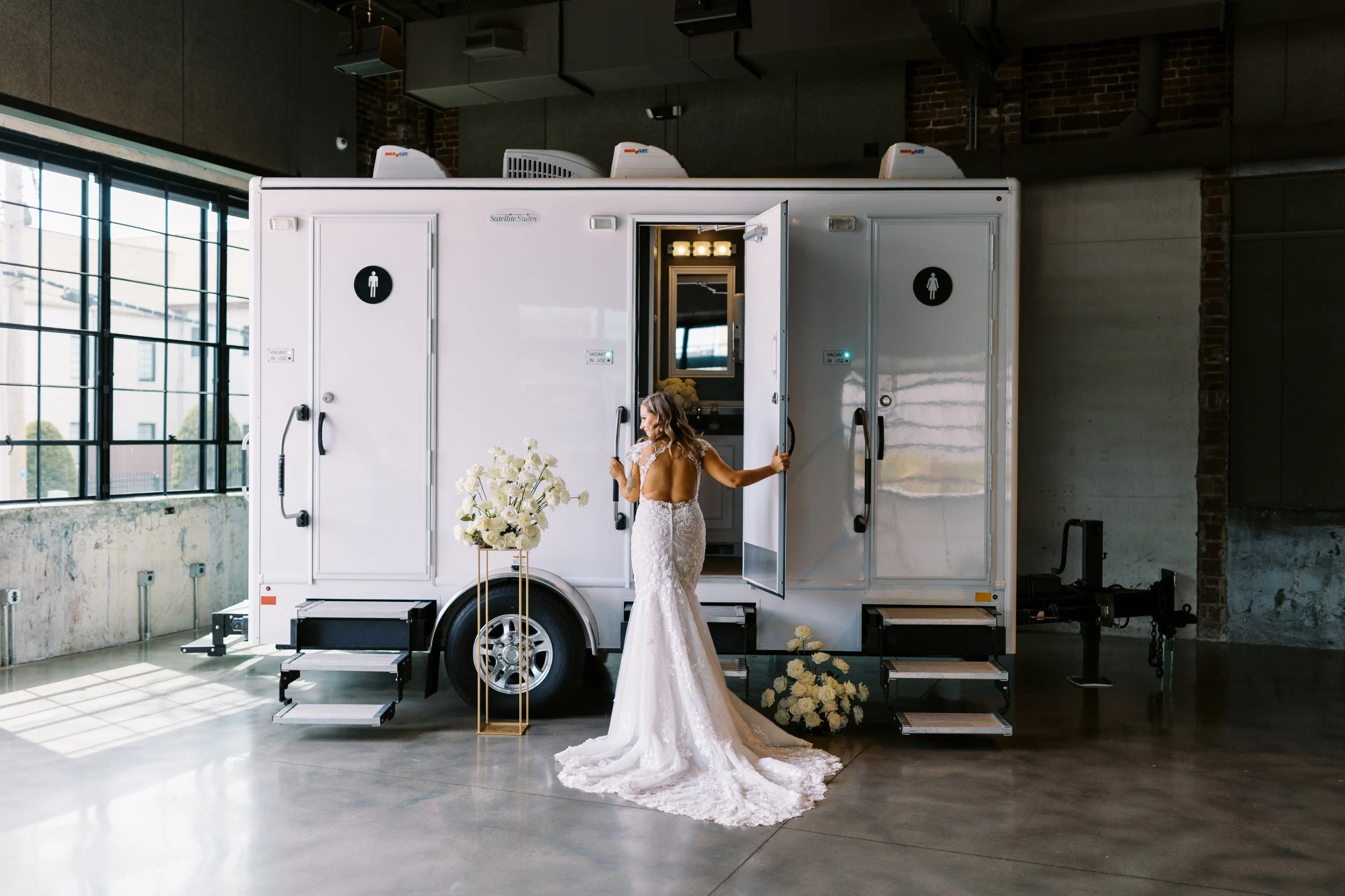 Bride in a wedding dress entering a mobile restroom trailer in an industrial-style building with large windows and exposed brick walls.