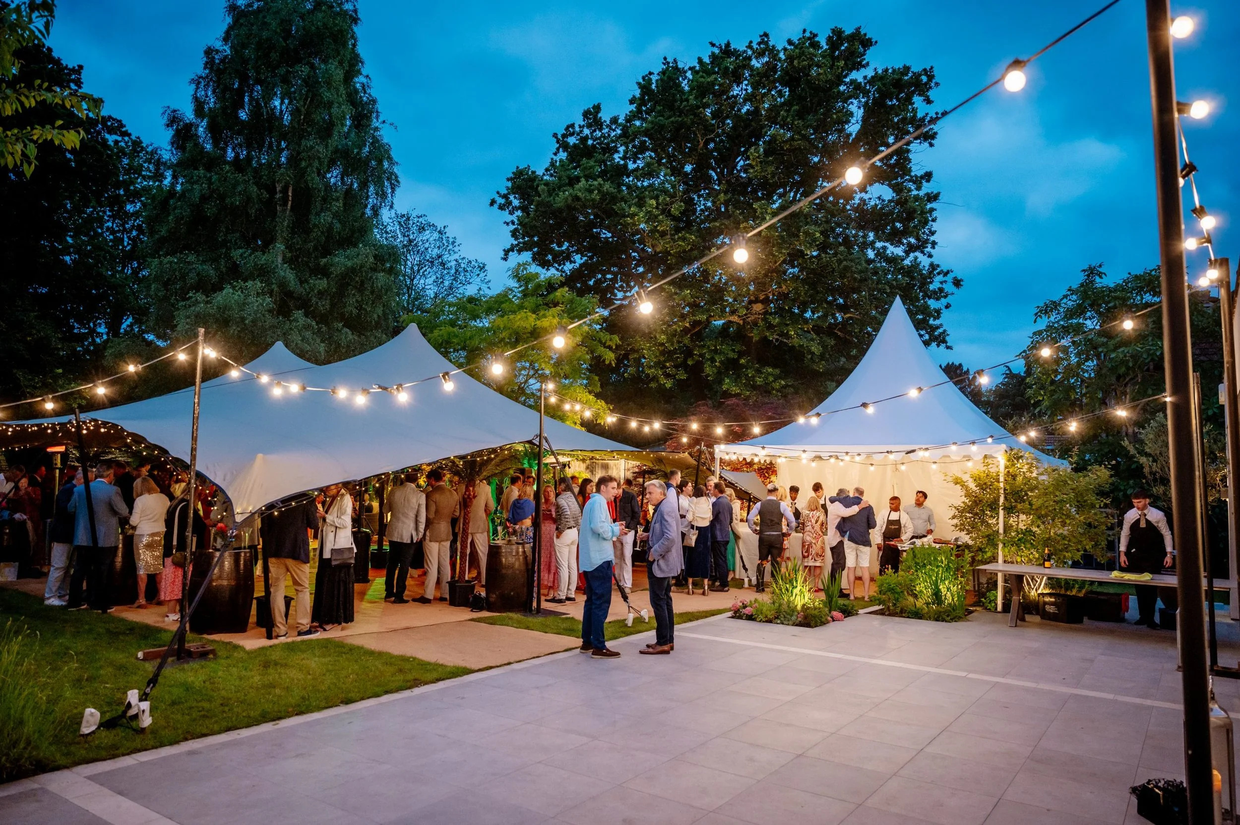 An outdoor evening event or party under large white tents with string lights, people mingling, and a garden area with plants and trees.