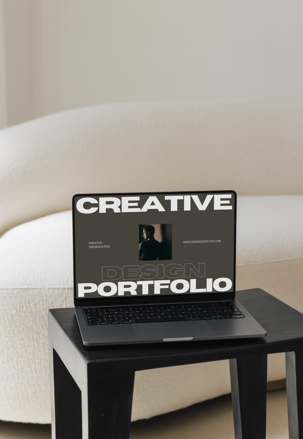 Laptop on a black table displaying a design portfolio cover with the word 'CREATIVE' and a photo of a man, in a room with beige furniture.