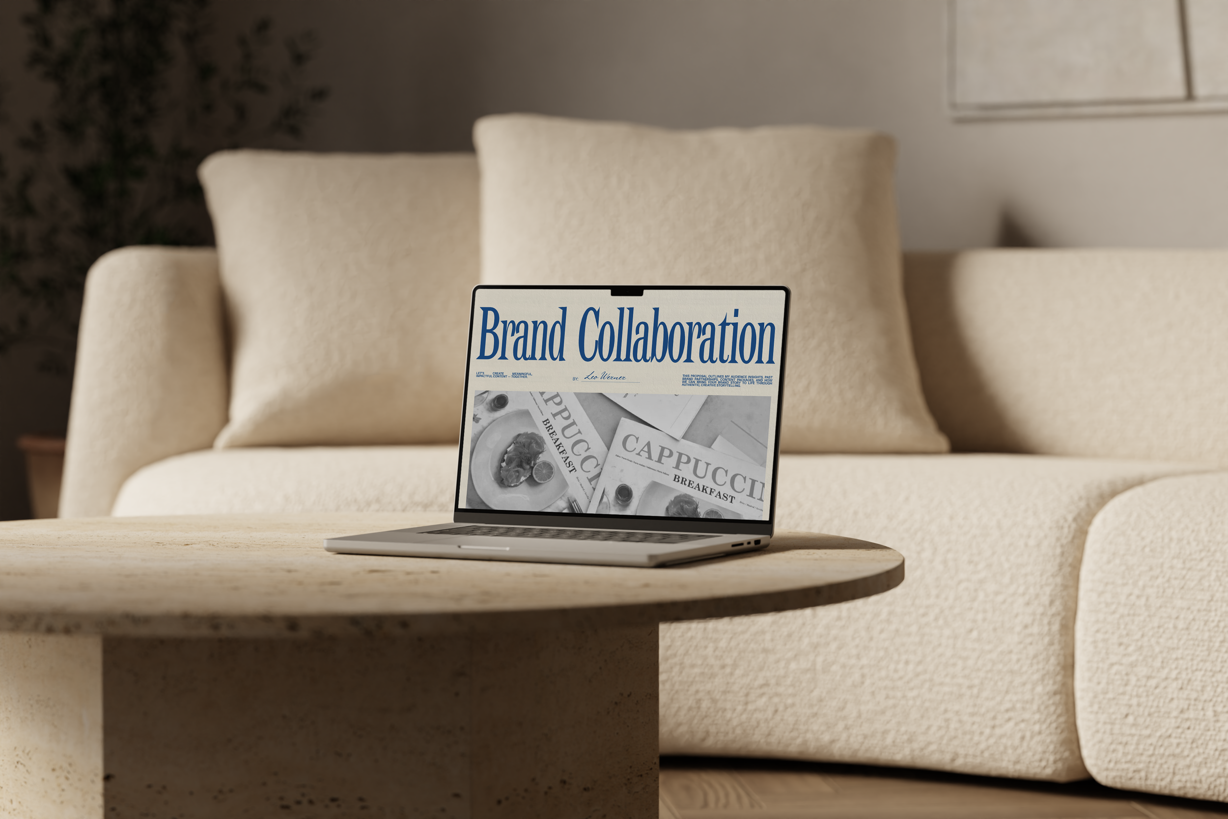A laptop on a wooden table displaying a magazine cover titled 'Brand Collaboration' with an image of dishes labeled 'Cappucci' and 'Breakfast' in a bright living room with a cream sofa and pillows.