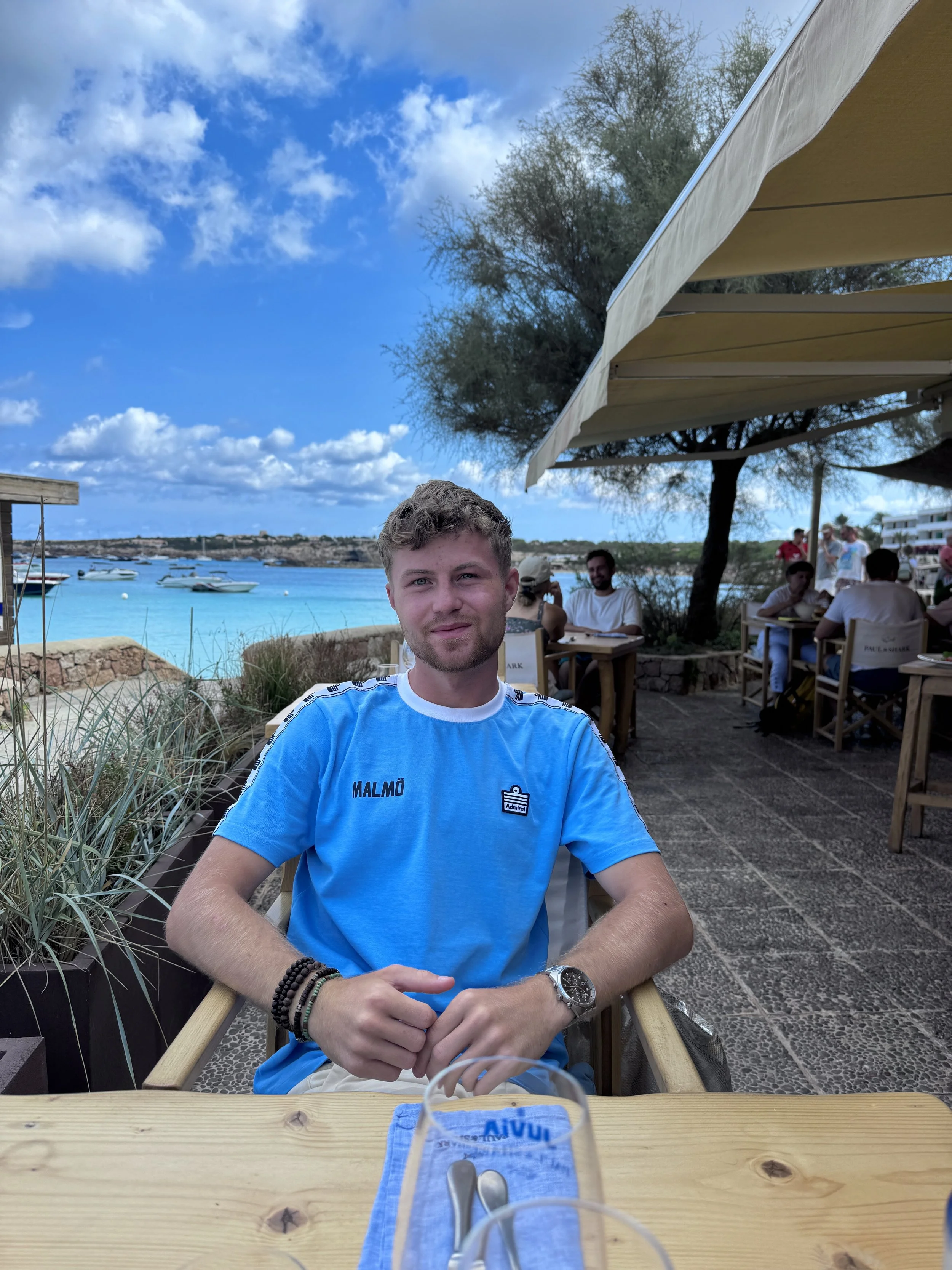 A young man with light skin and short light brown hair sitting at a table outdoors at a seaside restaurant, with a view of boats on the water and a partly cloudy blue sky.