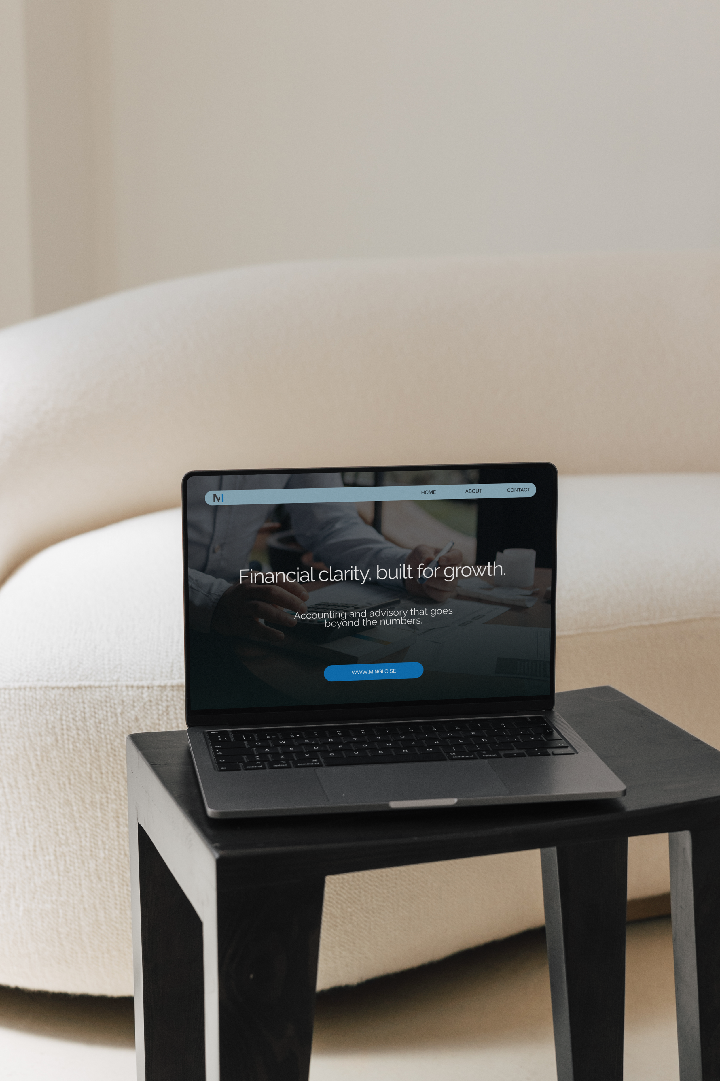 A laptop on a small black table displays a finance website with the headline 'Financial clarity, built for growth.' and navigation options for Home, About, and Contact. The background shows a cream-colored sofa.