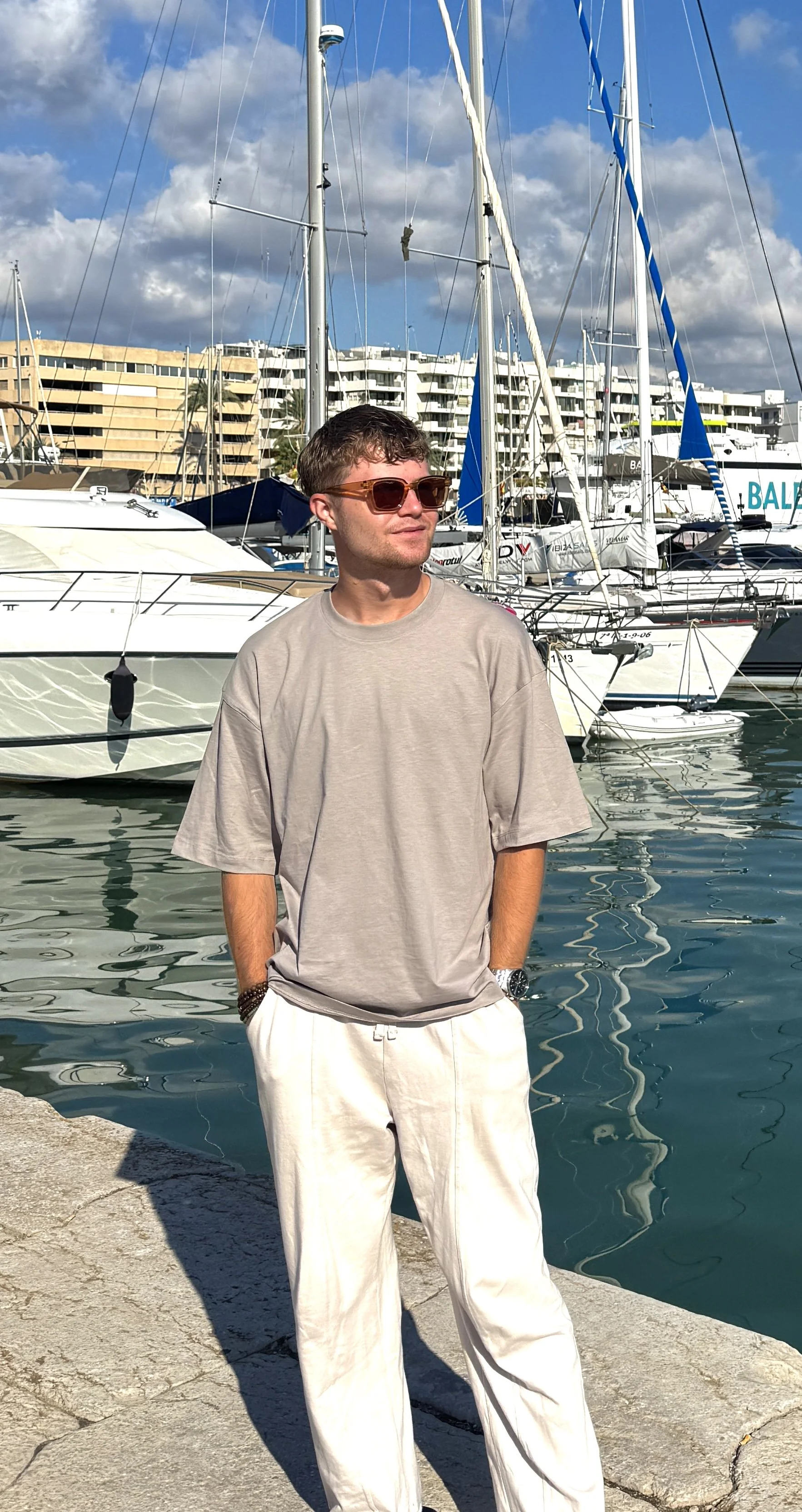 A man wearing sunglasses, a beige t-shirt, and white pants standing on a stone dock with sailboats and a marina behind him, under a partly cloudy sky.