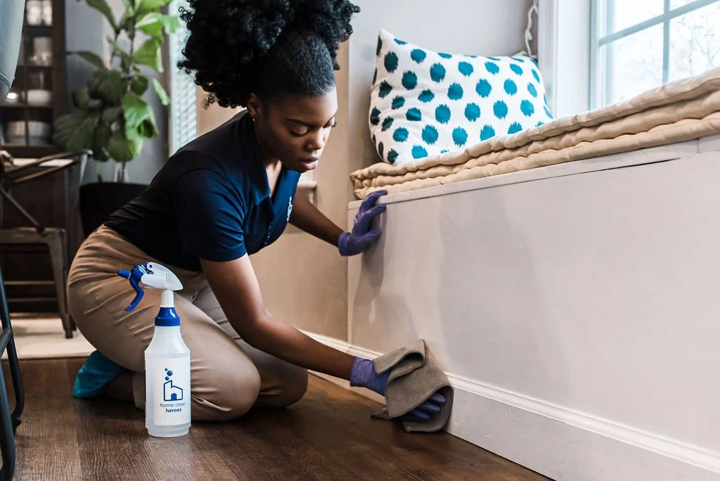 A woman wearing purple gloves is cleaning the baseboard of a bed with a cloth. She has a spray bottle labeled 'home clean heroes' placed on the wooden floor nearby. The bed has a blue and white polka dot pillow and layers of beige blankets, near a window with white curtains.
