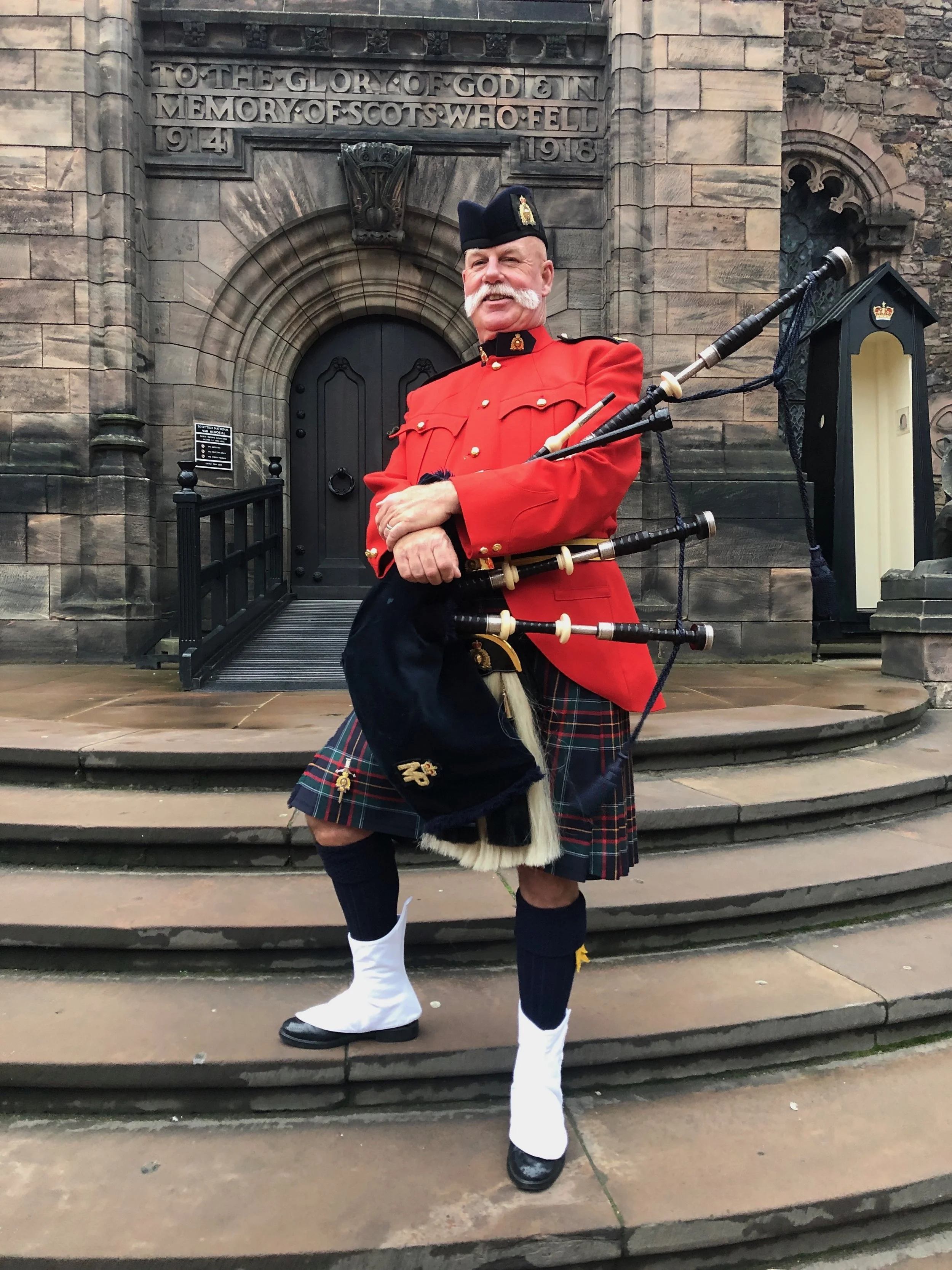 A man in traditional Scottish military attire, including a red jacket, kilt, and sporran, standing on stone steps in front of a stone building with a plaque and an arched doorway.
