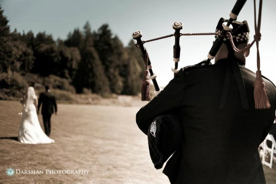 A photographer capturing a wedding scene on a beach, with a bride and groom walking in the background and a clear sky.