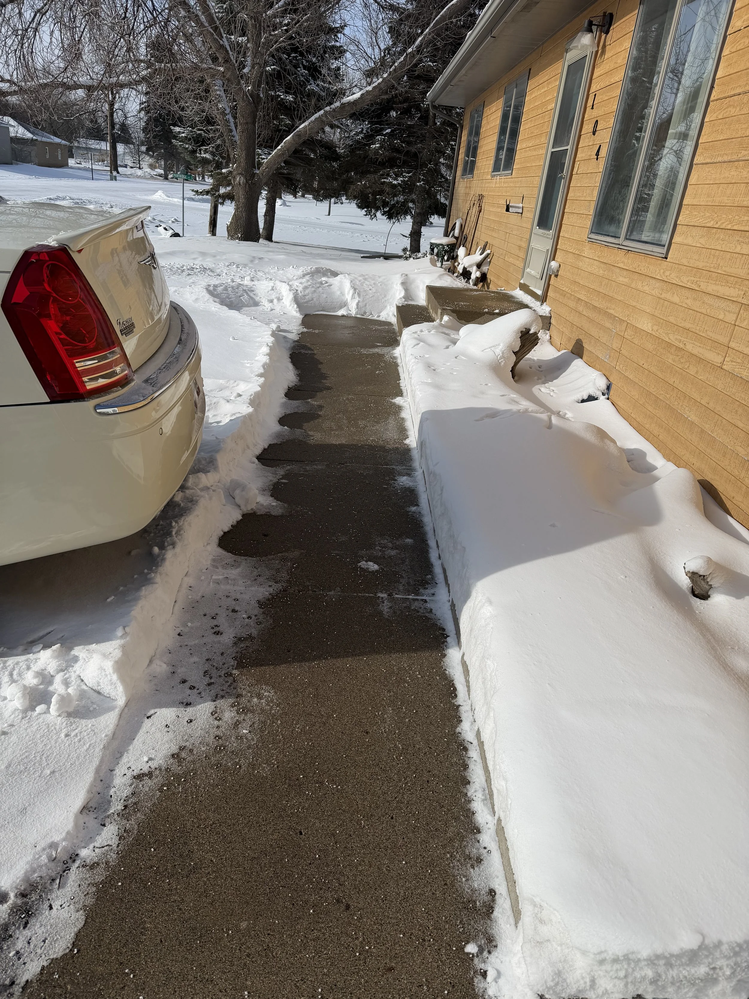 Snow-covered sidewalk and porch next to a house with yellow siding, after a snowfall, with a parked car on the left and leafless trees in the background.