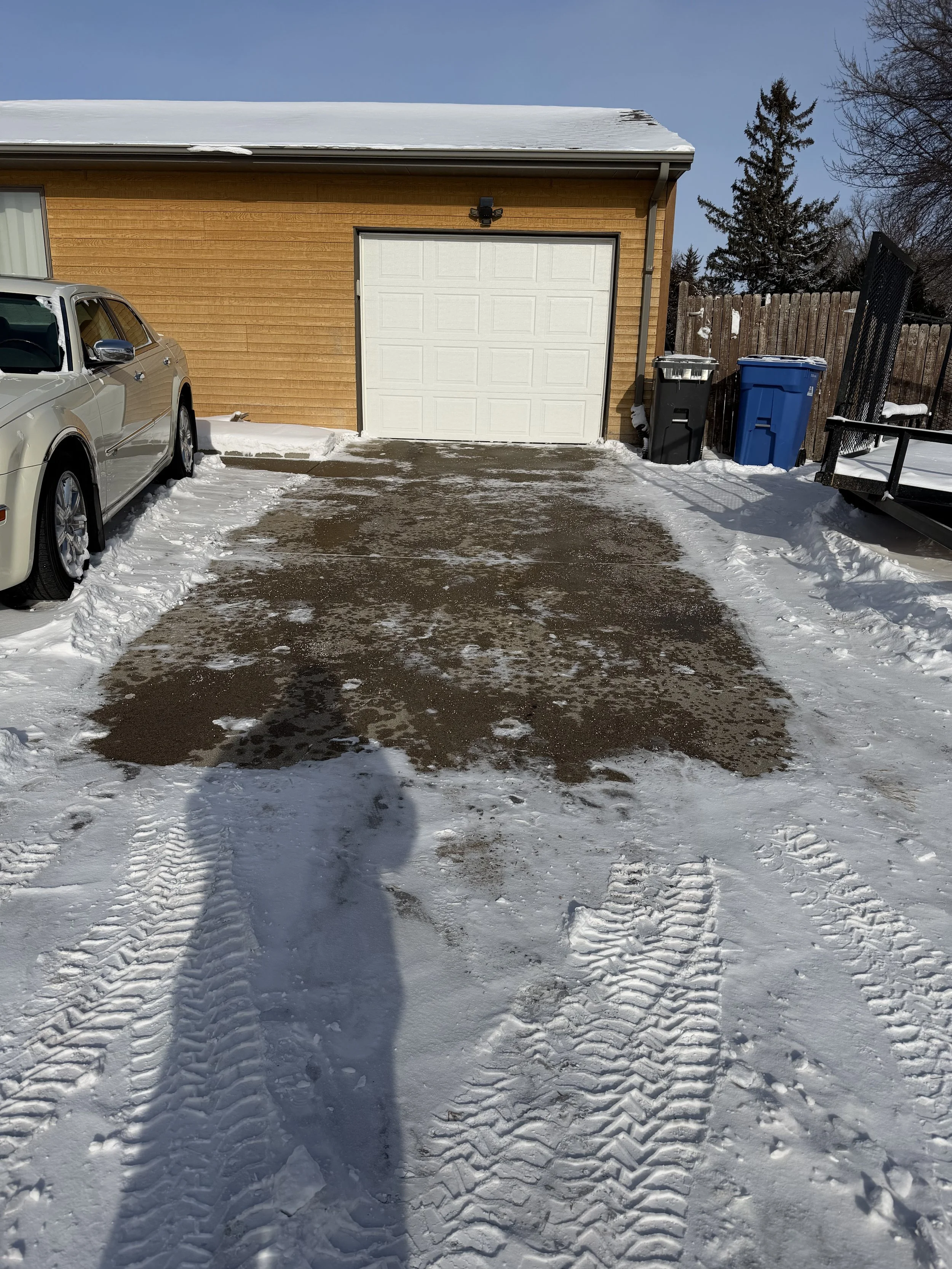 Snow-covered driveway with patches of cleared pavement leading to a closed garage door. A silver car is parked on the left side, and two trash bins, one black and one blue, are on the right side near a wooden fence. Tire tracks and footprints are vis