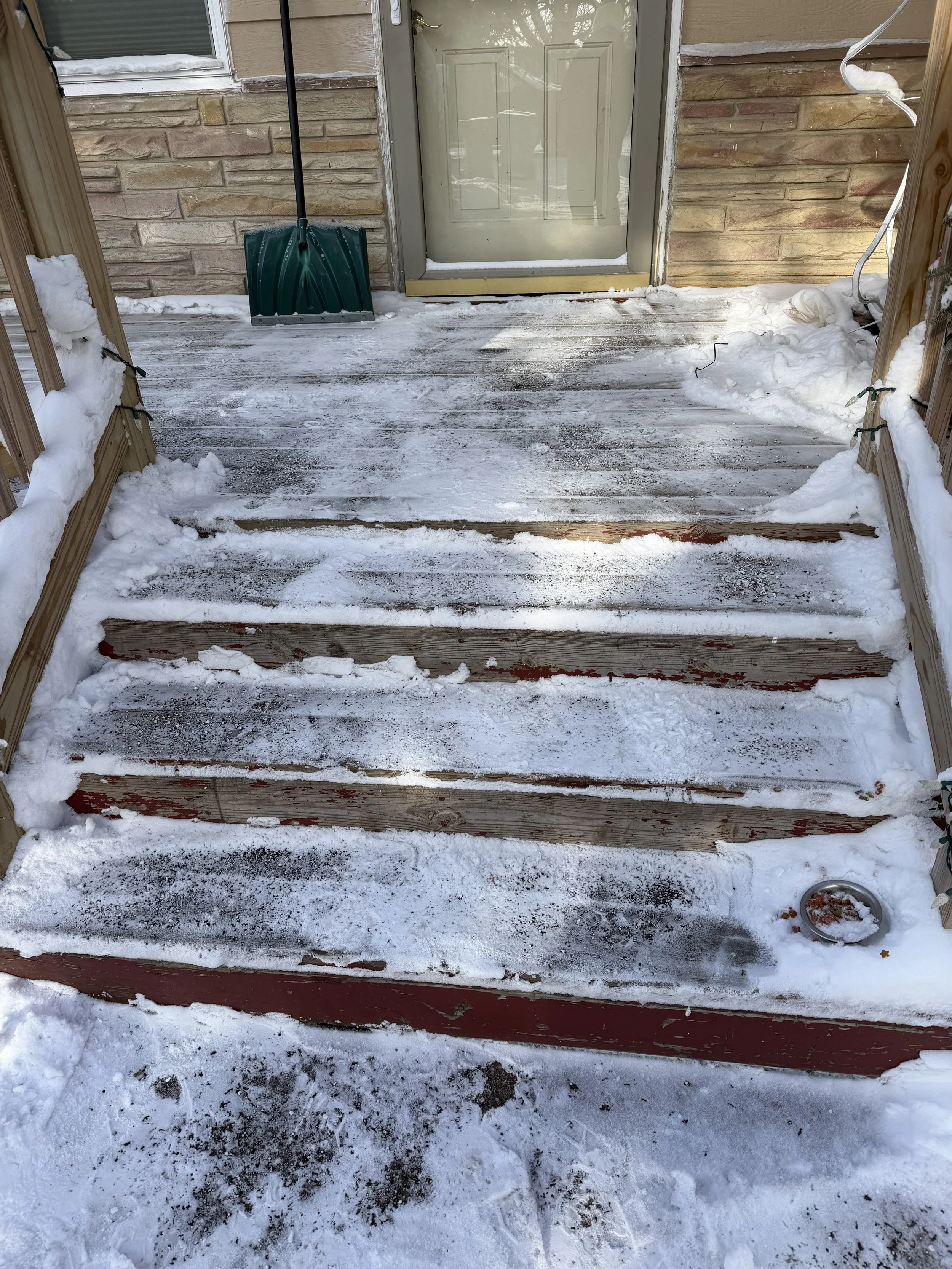 Snow-covered front porch with three wooden steps, ice, snow, and footprints. Green snow shovel leans against the house near the door. Snow and power cords on the porch railing and near the door.