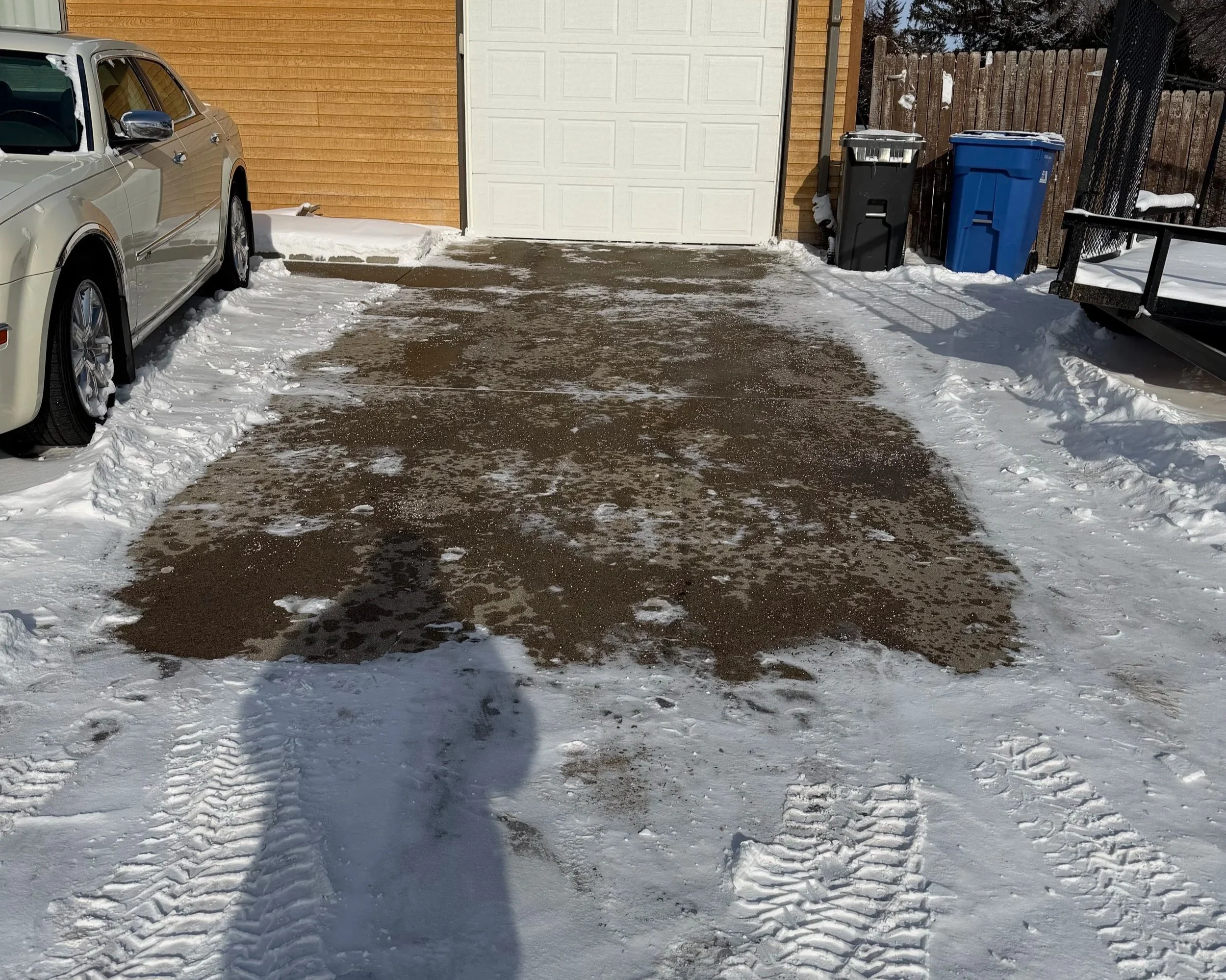 Snow-covered driveway with patches of cleared pavement, parked car on the left, two trash bins near a garage, a snow-covered deck on the right, and tire tracks in the snow.