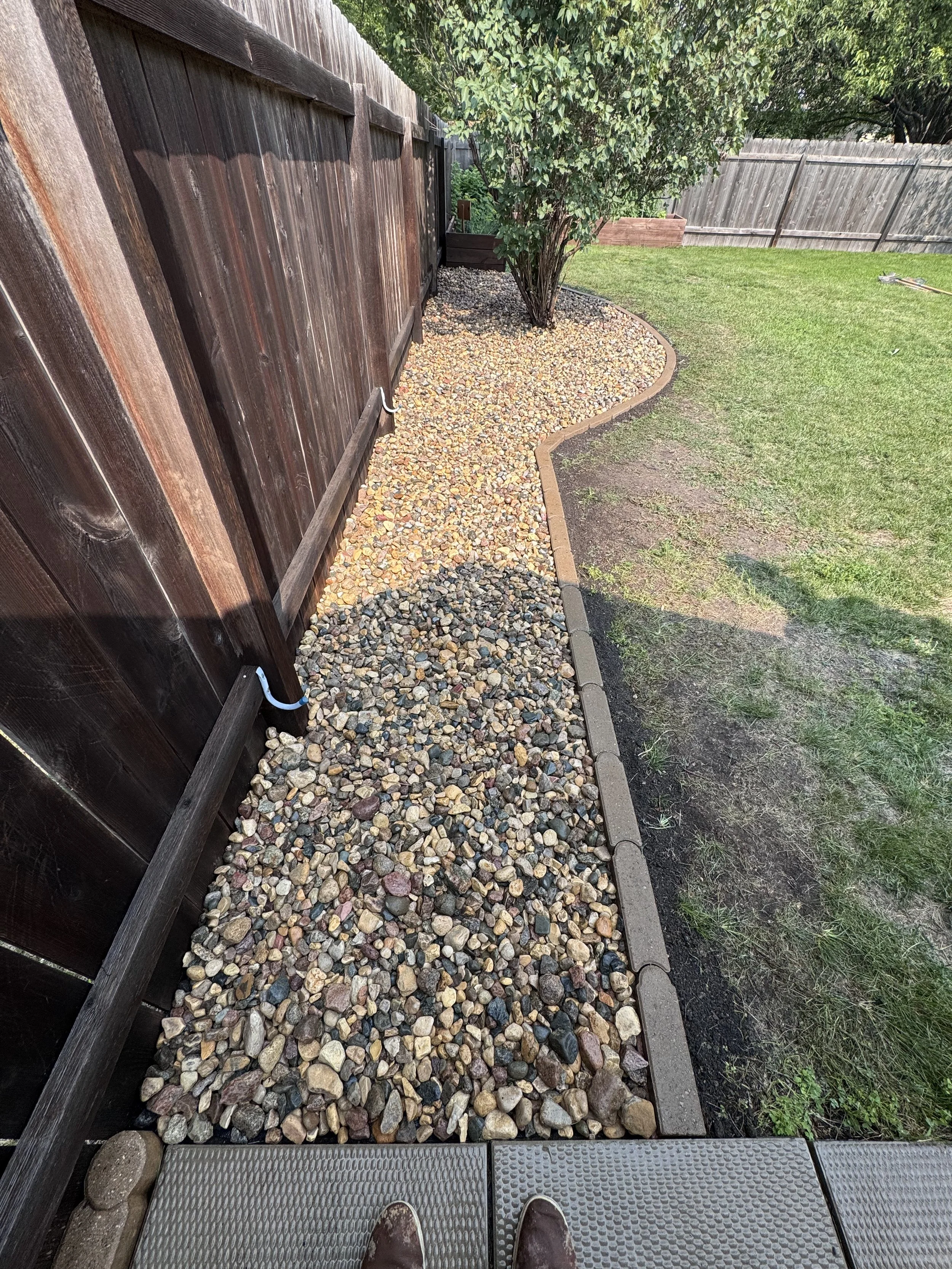 A backyard pathway made of loose multicolored gravel, bordered by a brick edge, with a wooden fence on the left and a grassy yard on the right, along with a tree and a raised garden bed in the background.