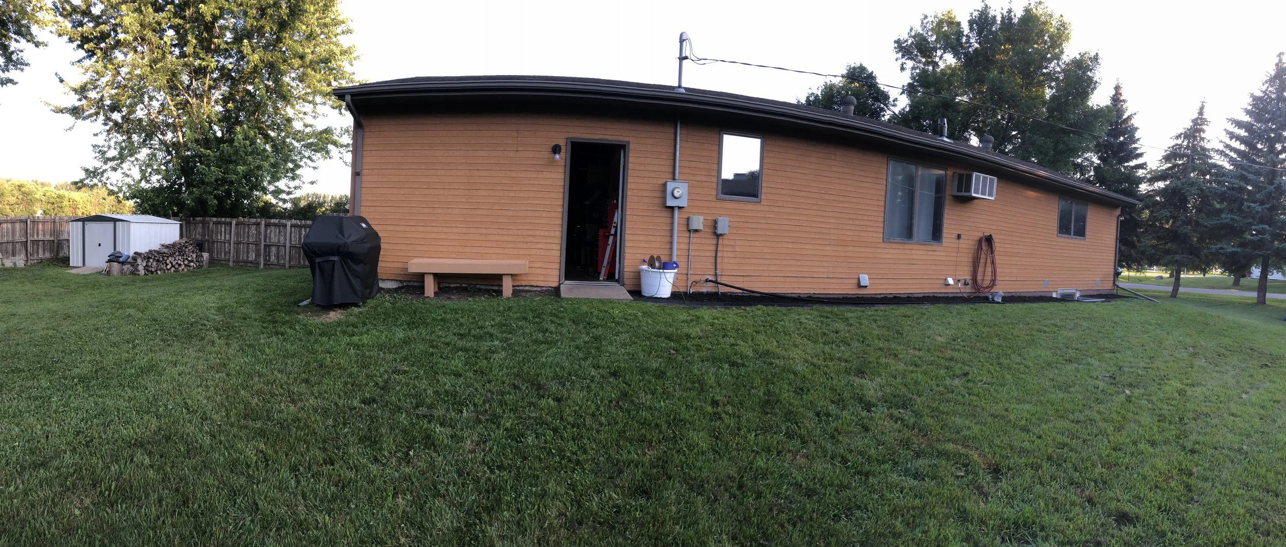 Backyard view of a house with tan wooden siding, a small door, electricity meters, and a window air conditioner. The yard has green grass, a covered grill, woodpile, and a shed. Trees and a wooden fence are in the background.