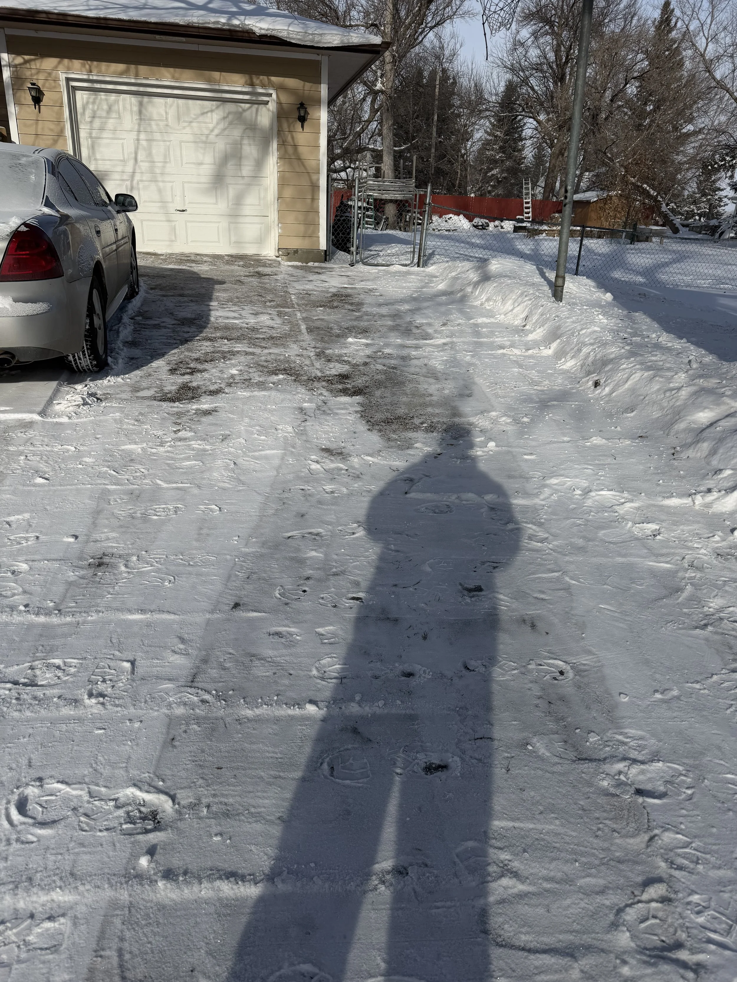 Snow-covered driveway with footprints, a parked silver car on the left, a house with a garage door in the background, and a chain-link fence with gate and snow on the ground.