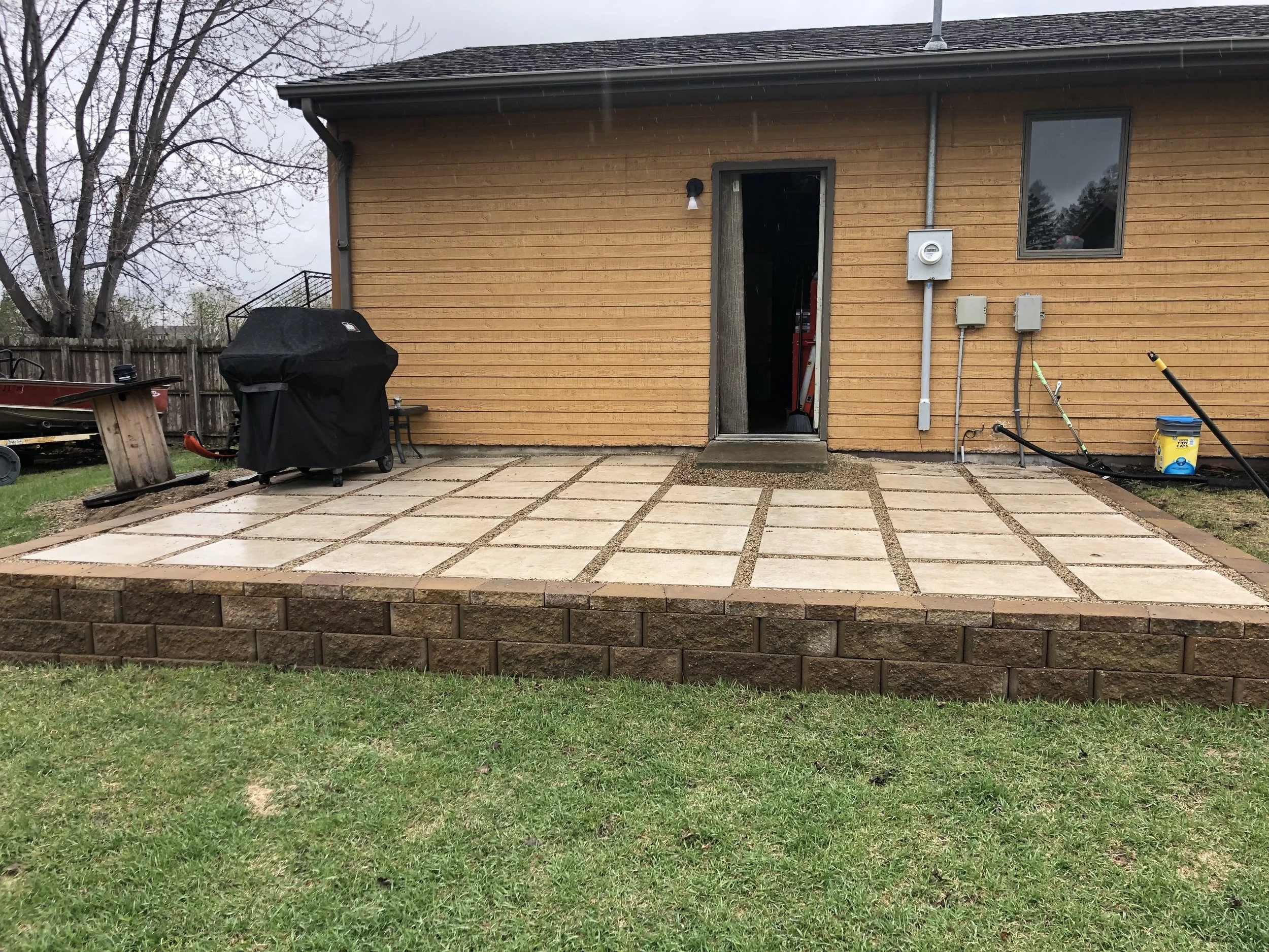 Newly constructed concrete patio with stone border in backyard, with outdoor grill and gardening tools near house with wooden siding.