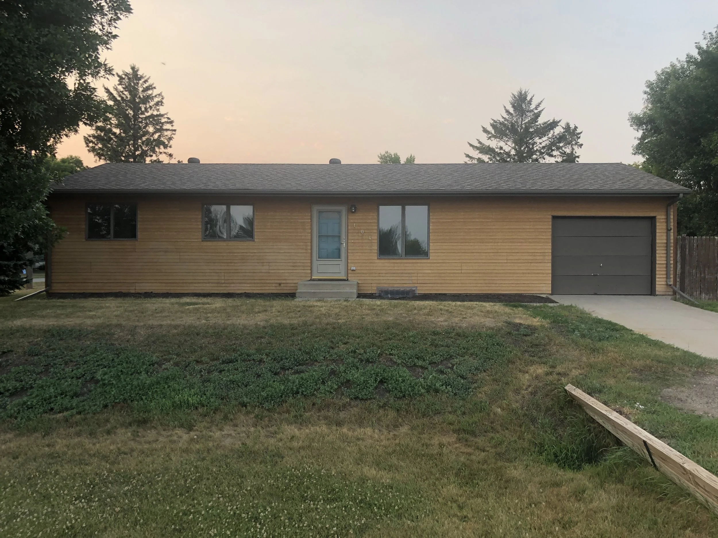 A single-story brick house with a gray garage door, a front door with stairs, two windows, and a front yard with grass and some plants, during sunset.