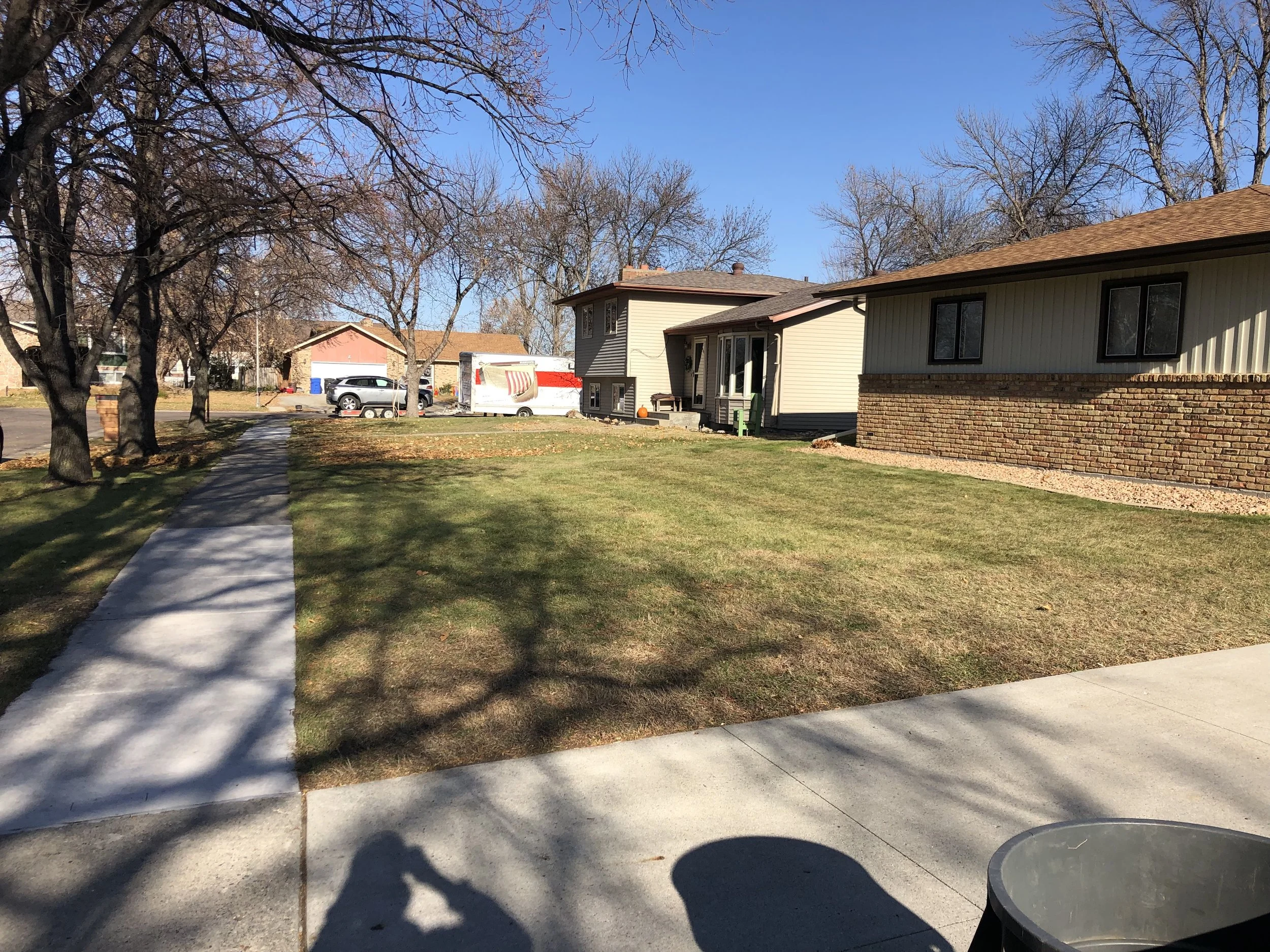 Suburban neighborhood with leafless trees, a sidewalk, and houses with yards. The scene is sunny with clear blue skies.